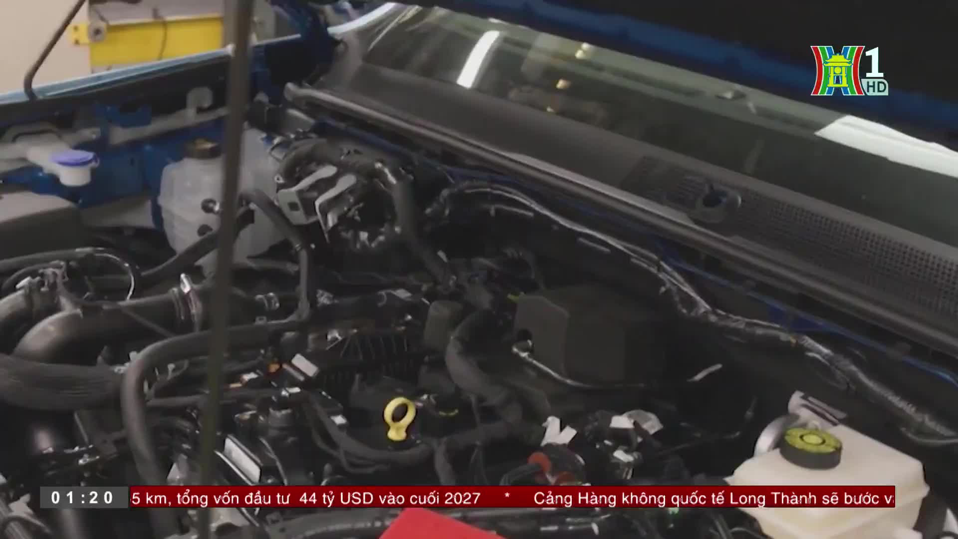 The open hood reveals the intricate engine of a blue vehicle, likely part of a segment from HanoiTV1. A yellow cap and various black tubes and wires are visible, suggesting a focus on automotive technology.
