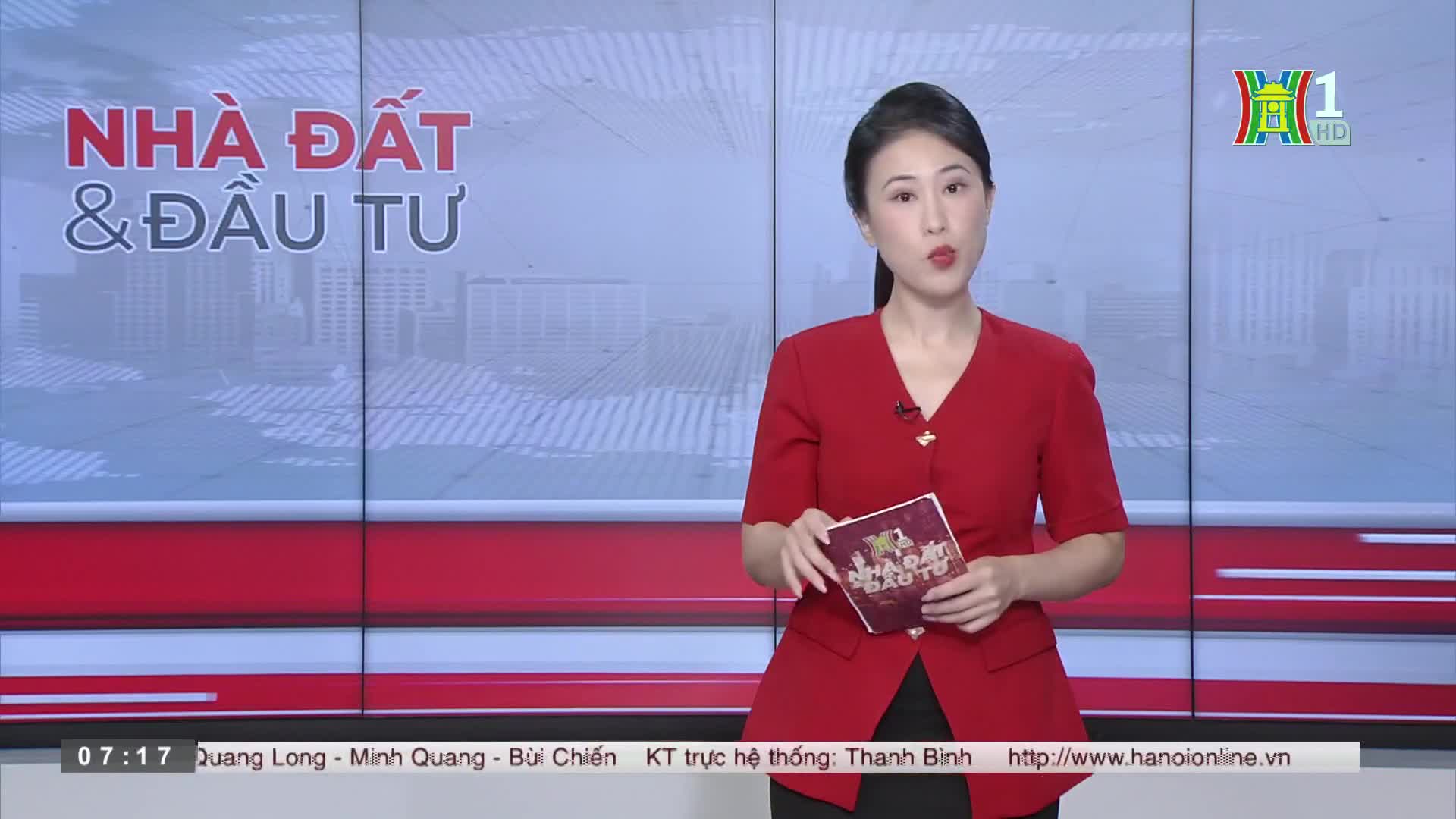 A woman in a red jacket stands in front of a backdrop, holding a small booklet. The HanoiTV1 logo is visible in the corner, and the screen behind her displays the words "NHÀ ĐẤT & ĐẦU TƯ".
