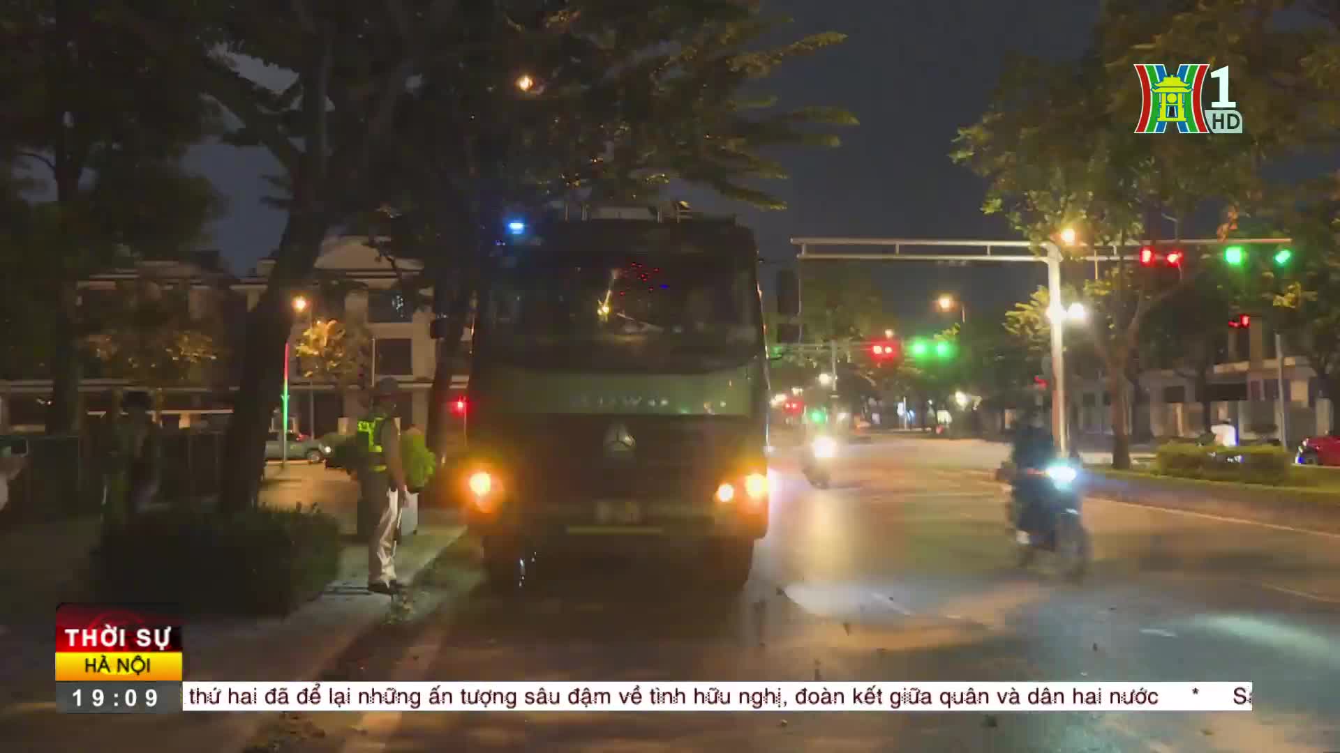 A large truck, its headlights on, is moving slowly down a street at night. The HanoiTV1 broadcast is showing a news report, with traffic lights visible in the distance.
