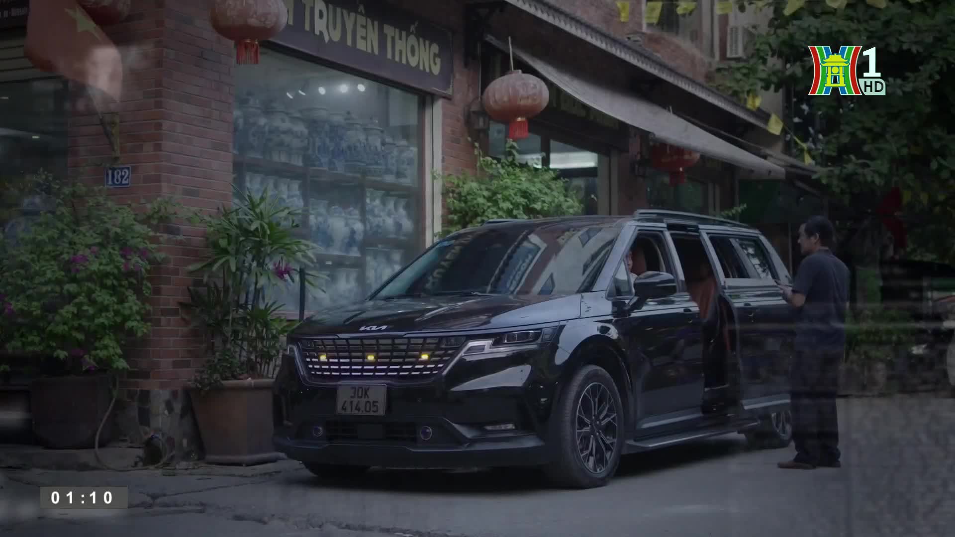 A black Kia minivan sits parked on a Hanoi street, its door open as a man stands nearby. The scene is framed by a shop with a sign in Vietnamese and a logo for HanoiTV1.

