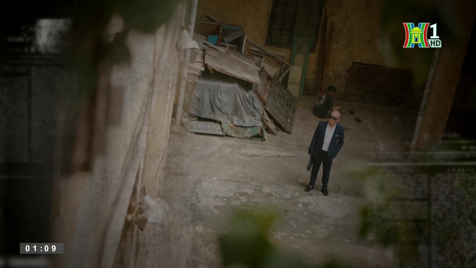 A man in a suit, holding something dark, stands in the middle of a concrete courtyard. A second person sits in the shadows near a pile of debris, as the HanoiTV1 logo appears in the corner.
