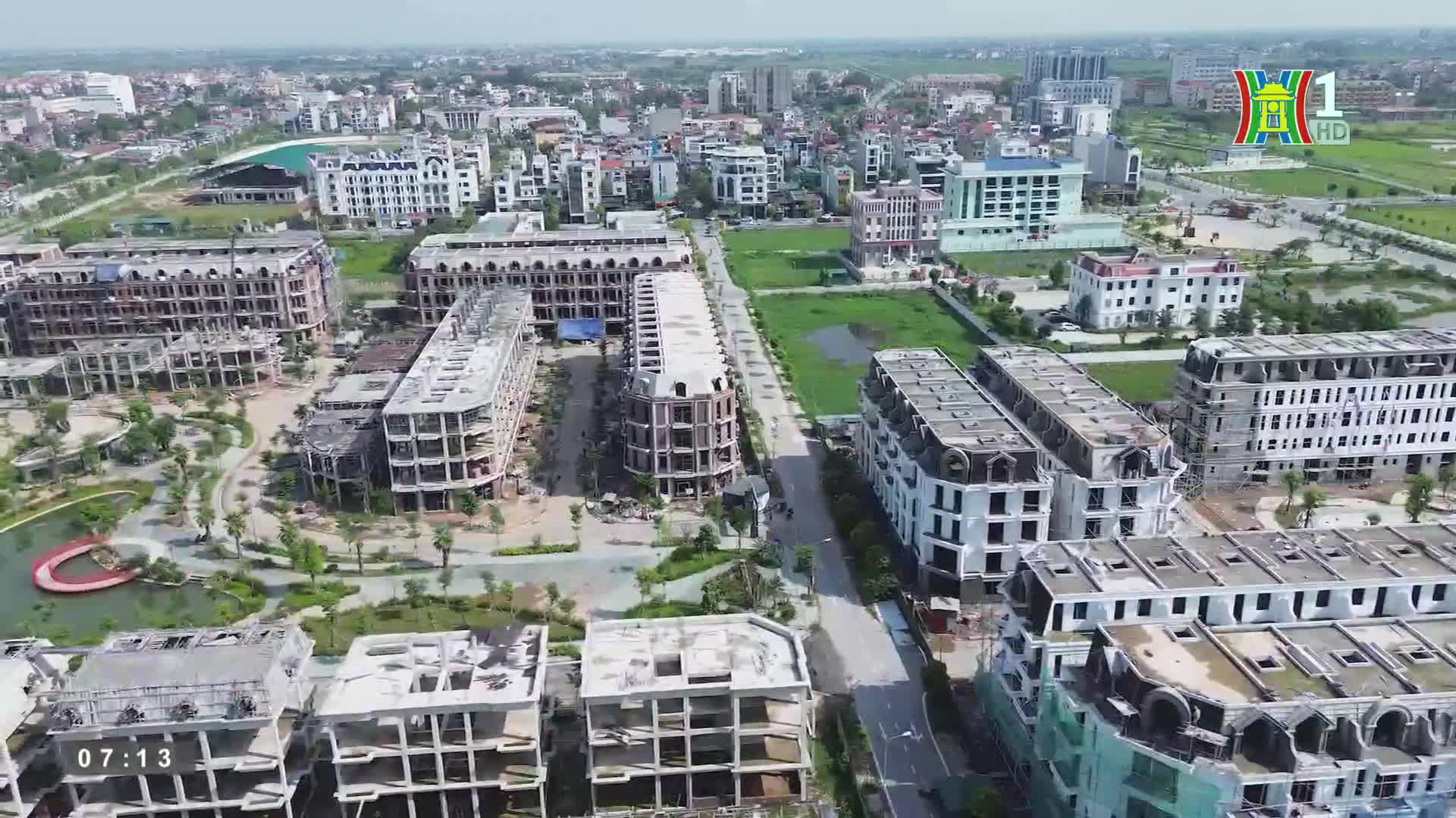 From HanoiTV1, a wide shot shows a new development in Vietnam, with rows of unfinished buildings lining a long road. The structures have white facades and dark roofs, and the surrounding area is mostly undeveloped.
