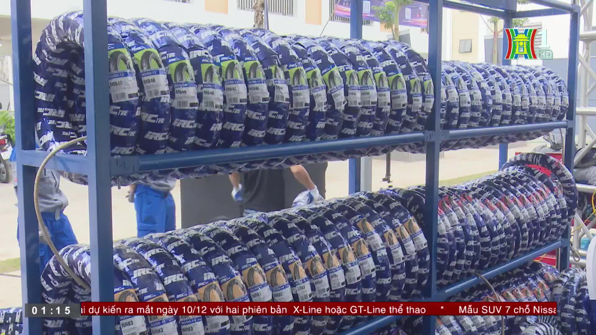Stacked tires, wrapped in blue and white, fill the metal shelving. A HanoiTV1 broadcast is likely showing the preparation for a new vehicle launch in Vietnam.
