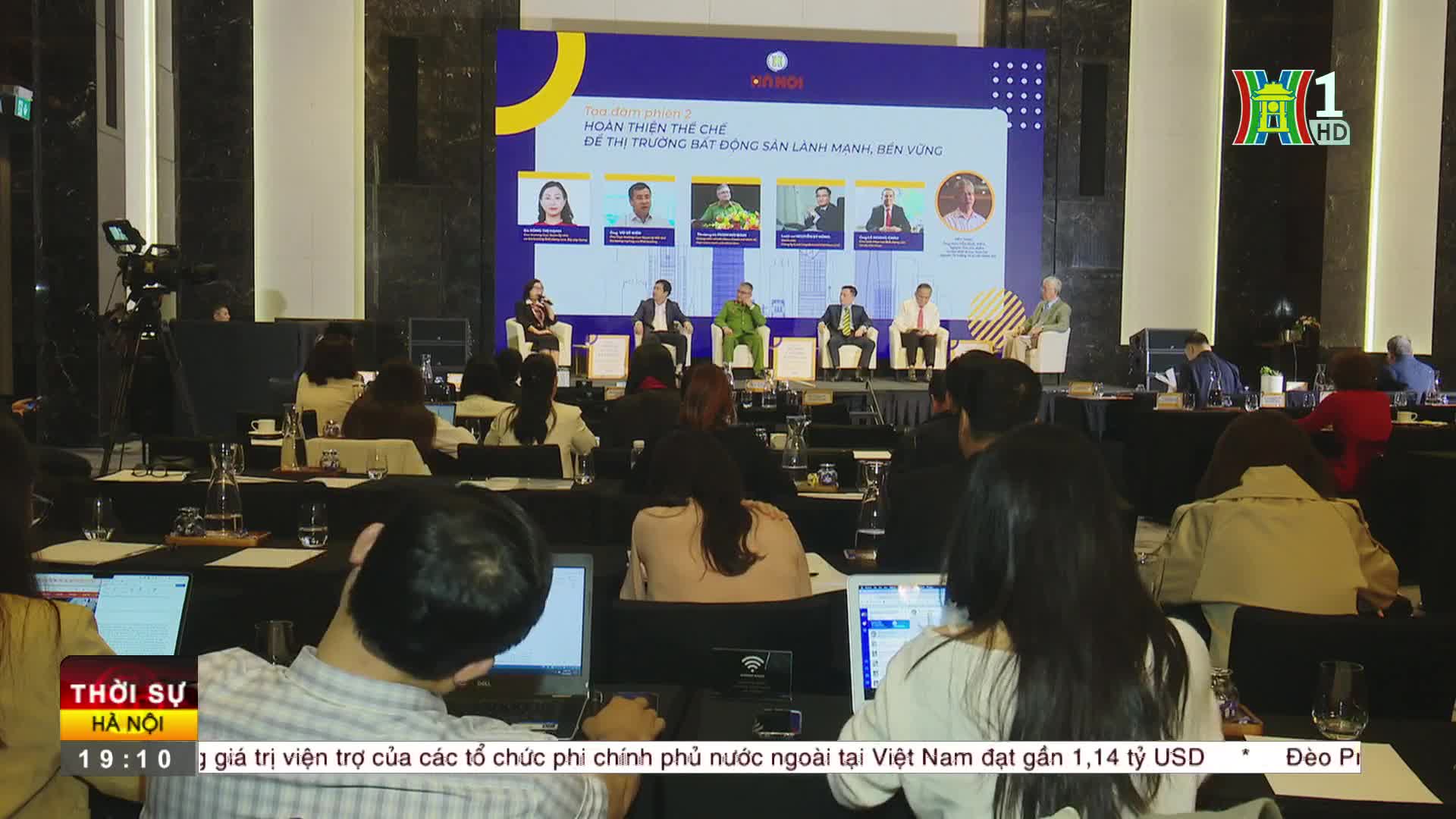On a stage, a panel of suited people sits beneath a banner, while an audience listens intently. HanoiTV1's logo is in the corner, and the bottom of the screen shows a news ticker.
