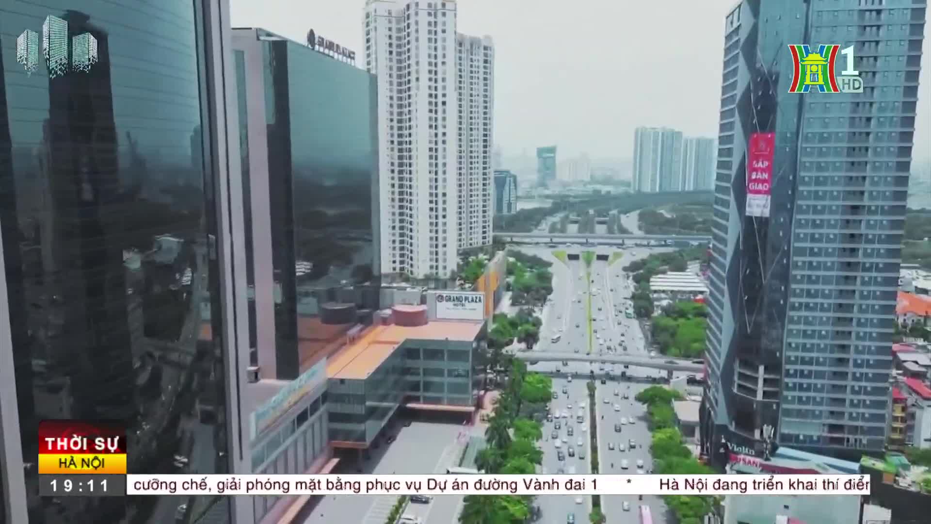 Traffic flows steadily along a wide, multi-lane road, flanked by tall buildings reflecting the overcast sky. The HanoiTV1 logo is visible in the corner, and the news ticker informs about a project on the Ring Road.
