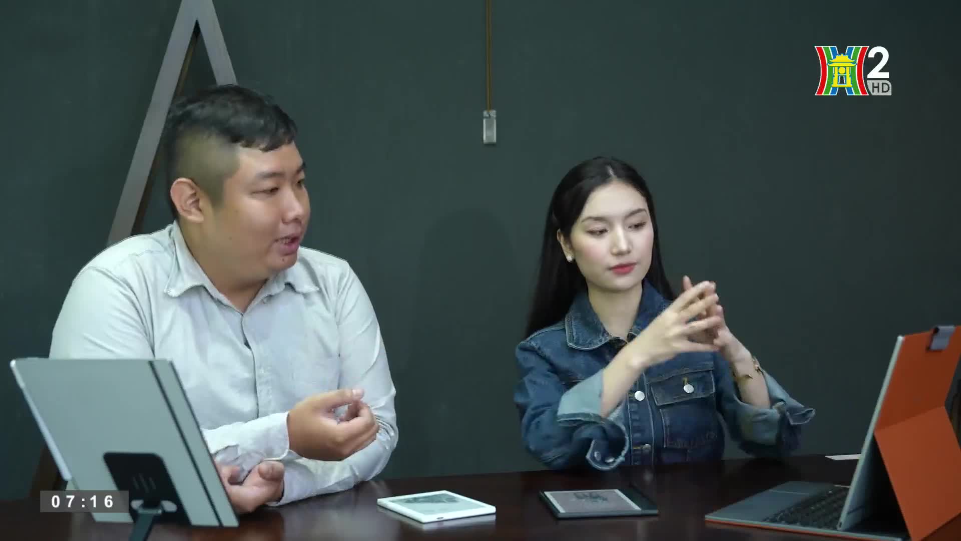 A man in a light shirt gestures with his hands as he speaks, while a woman in a denim jacket listens intently, her fingers clasped. The HanoiTV2 logo is visible in the background.
