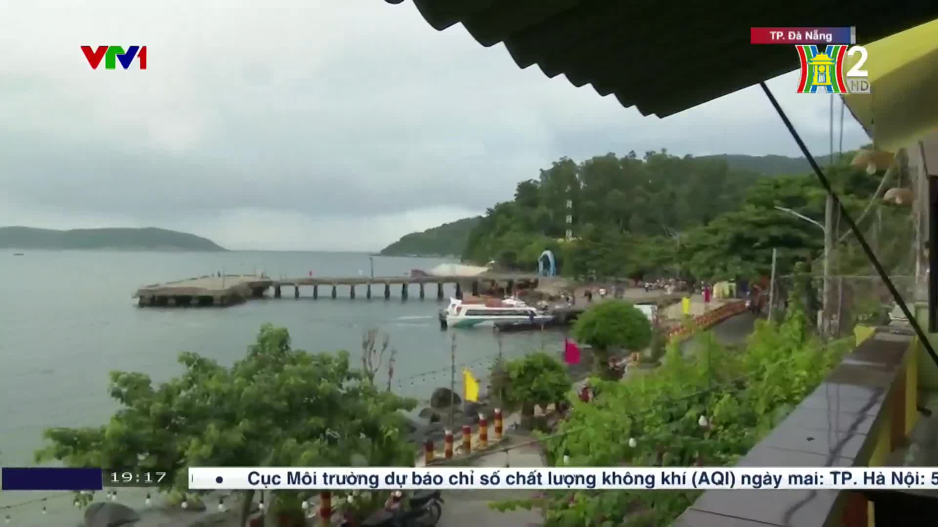 A white and blue boat is docked at a pier, with people milling about on the shore. The scene is being broadcast on HanoiTV2 from Da Nang in Vietnam, with a cloudy sky overhead.
