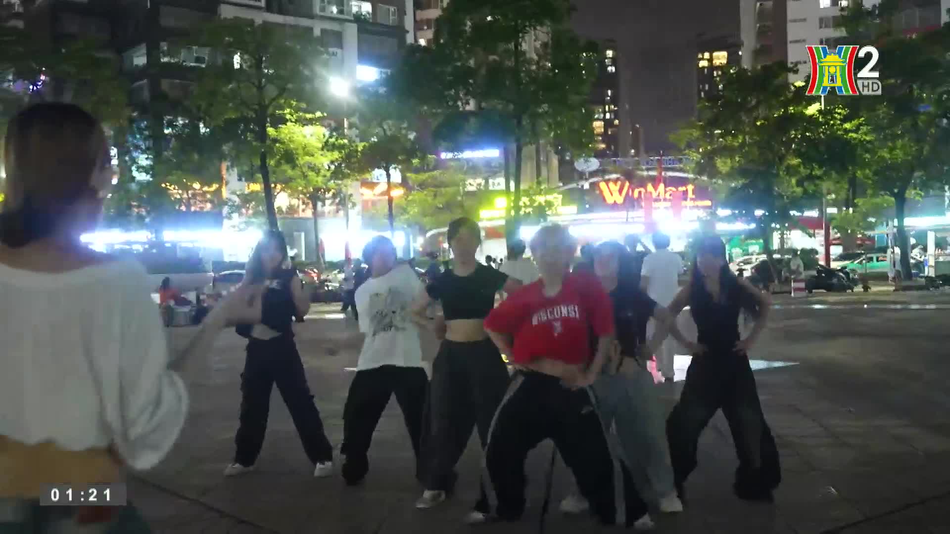 A group of young women are dancing on a plaza, their movements sharp and synchronized. The HanoiTV2 logo sits in the corner, broadcasting the scene as the dancers move in the evening light.

