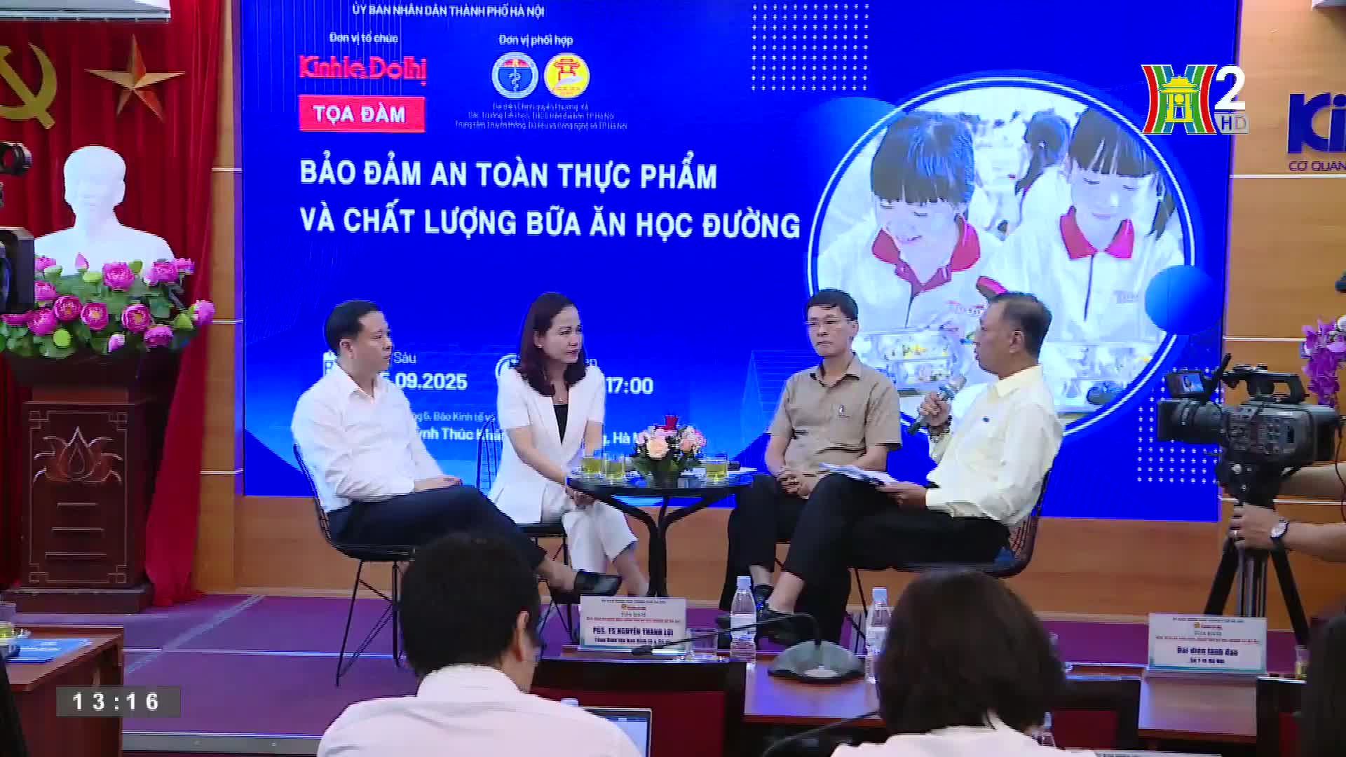 Four people sit on a stage, discussing food safety and school lunches, with a HanoiTV2 logo visible in the background. A camera operator films the panel, capturing the discussion for the broadcast.
