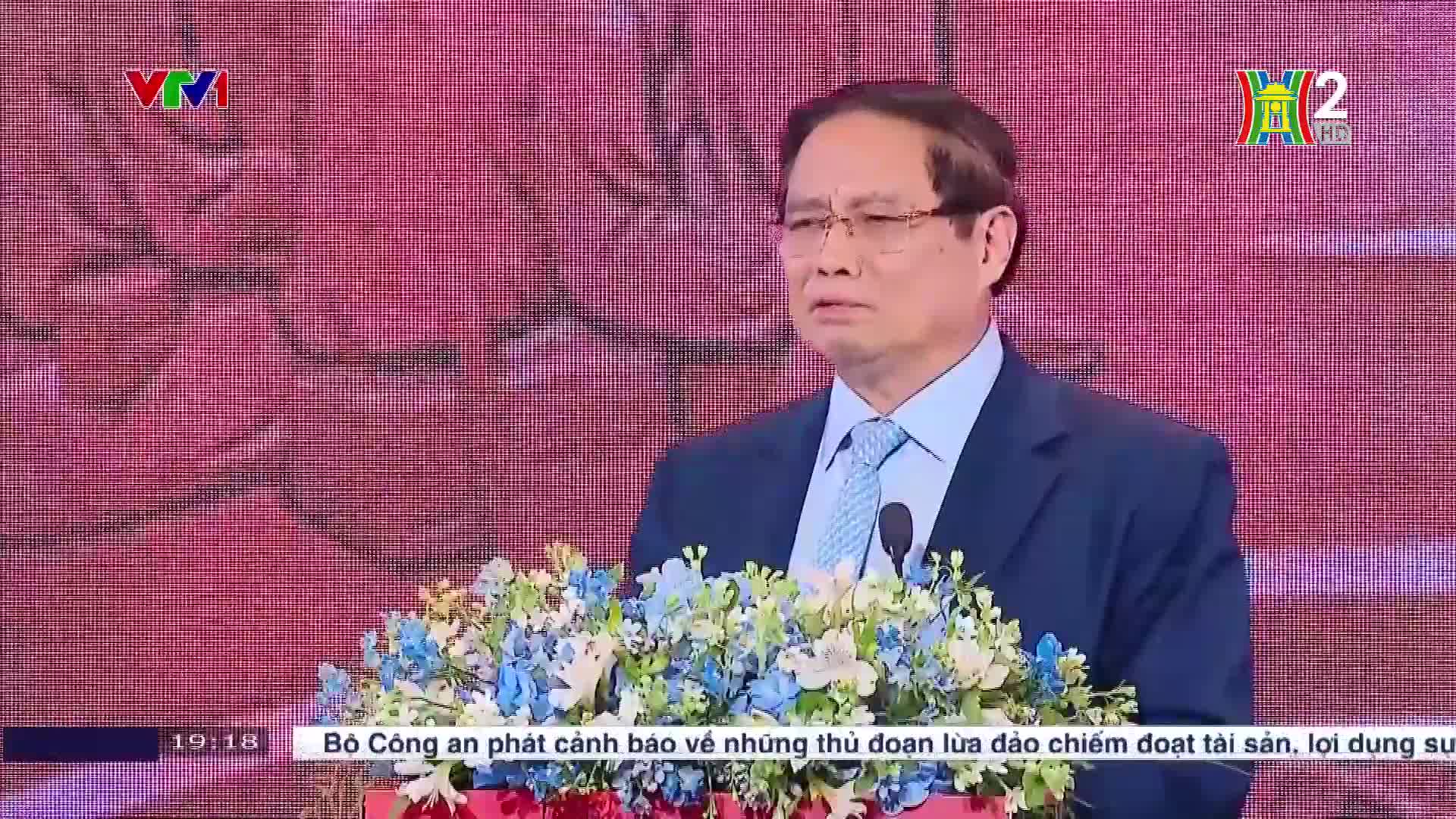 A man in a blue suit and tie speaks at a podium adorned with flowers, the Vietnam Television logo and HanoiTV2 logo are visible. The bottom of the screen displays a news ticker in Vietnamese.
