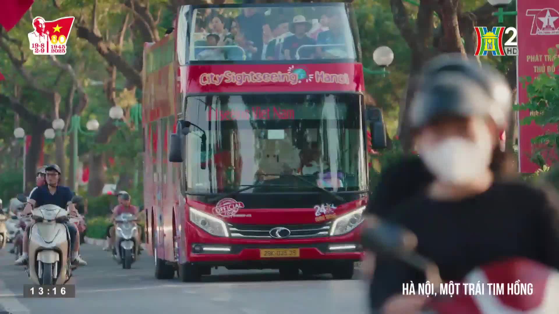 A bright red double-decker City Sightseeing bus rolls down the street, packed with tourists. Scooters weave around it, while a HanoiTV2 graphic and a banner celebrating Vietnam's history are visible in the background.
