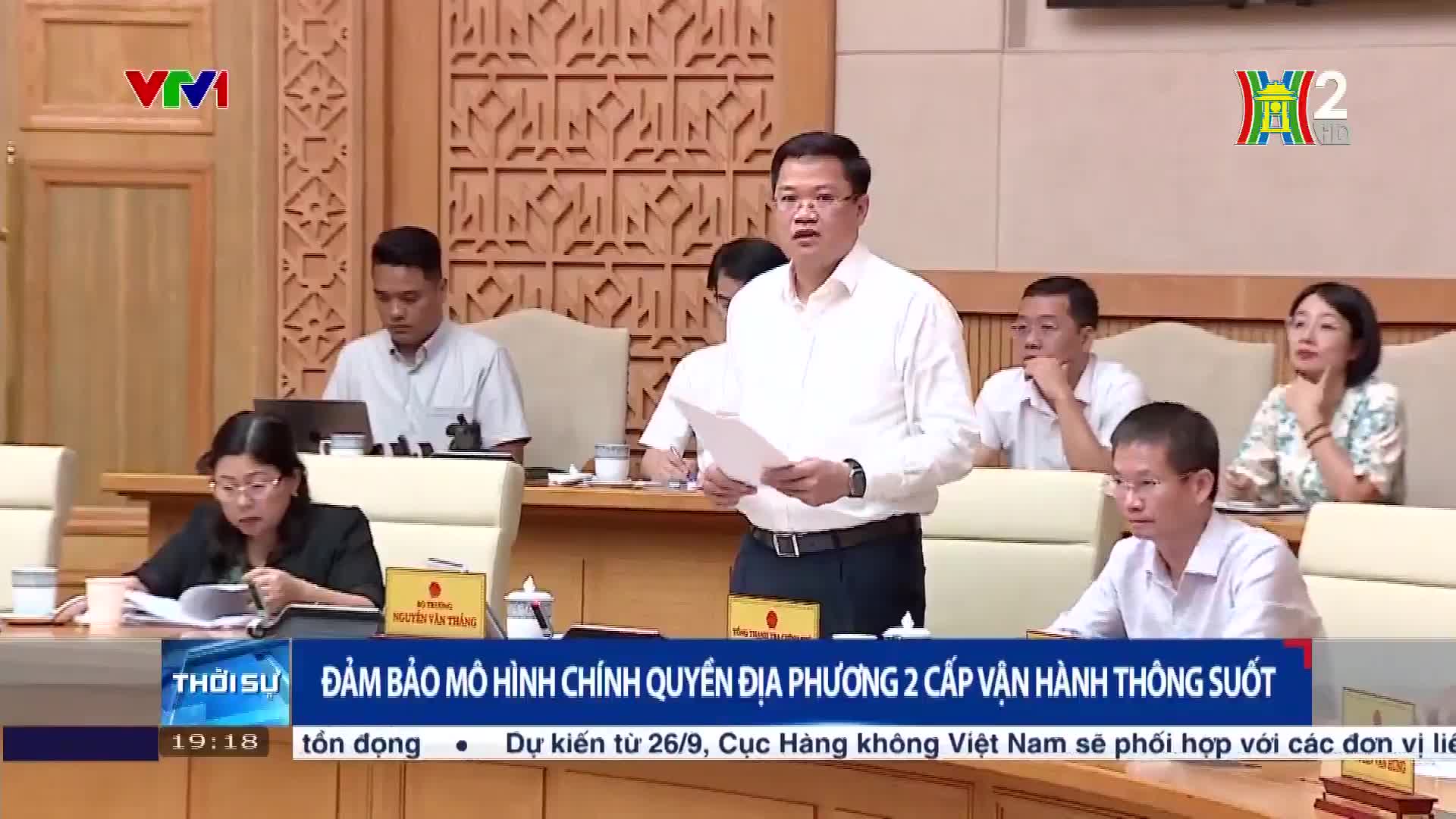 A man in a white shirt gestures while speaking, holding papers. He's standing in front of a panel of people, with a HanoiTV2 logo in the corner.
