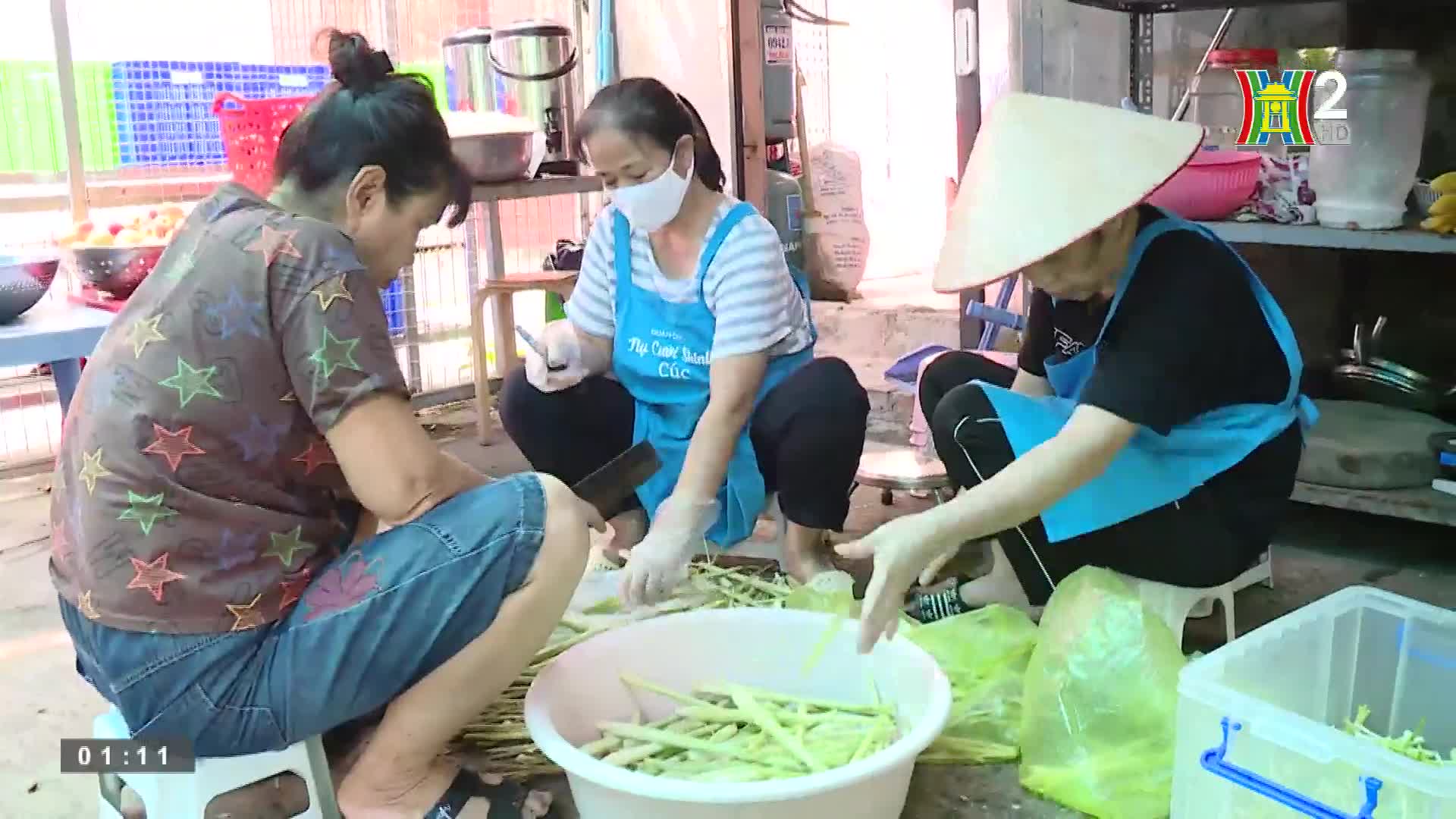 Three women, seated on small stools, are meticulously preparing vegetables. The woman in the center, wearing a mask, uses a cleaver, while the others sort and arrange the trimmed pieces.

