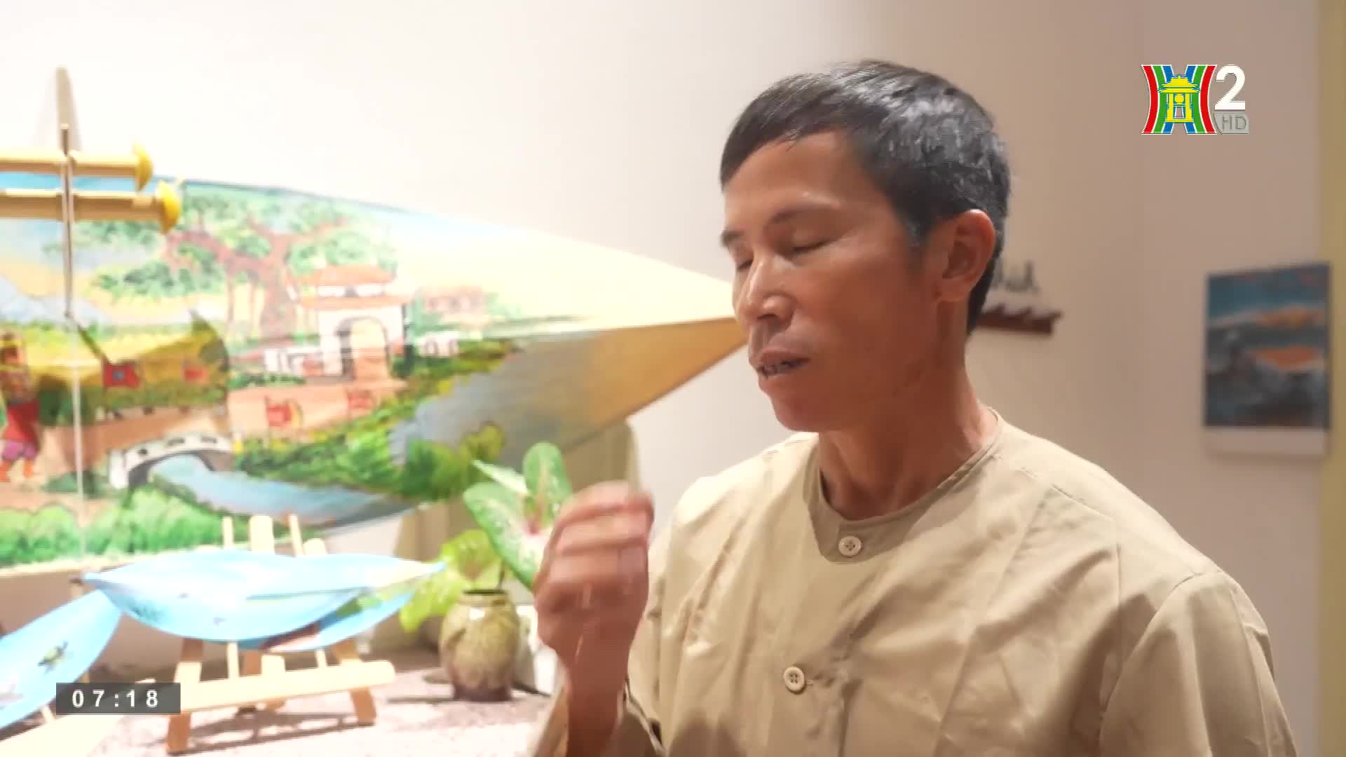A man in a light-colored shirt speaks, gesturing with his right hand. Behind him, a painting depicts a landscape with a bridge and trees, likely part of a segment on HanoiTV2.
