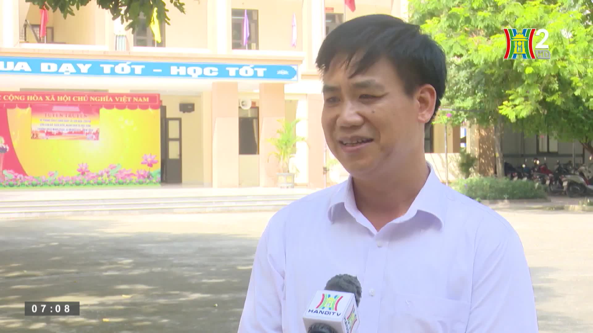 A man in a white shirt speaks into a microphone bearing the HanoiTV2 logo. Behind him, a banner with Vietnamese text sits in front of a school building.
