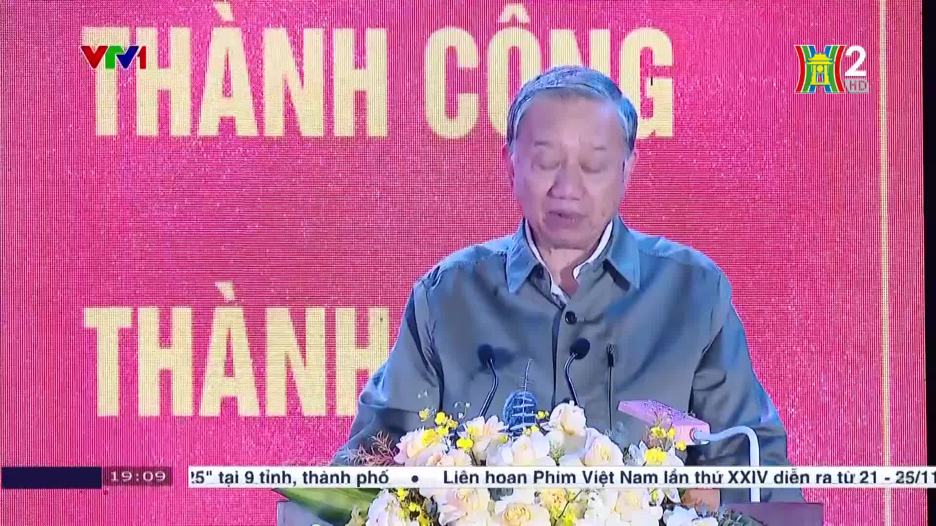 A man in a green shirt is speaking at a podium, with the Vietnam Television logo visible on the screen behind him. The broadcast is from HanoiTV2, and the background is a bright pink with the words "Thanh Cong" in large white letters.
