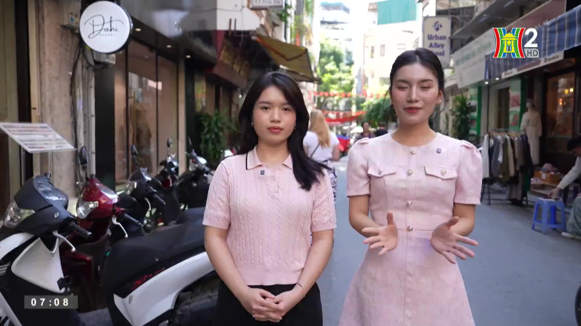 Two women in matching pink outfits stand on a narrow street, gesturing with their hands. Behind them, motorbikes line the sidewalk, and shops and pedestrians fill the background.
