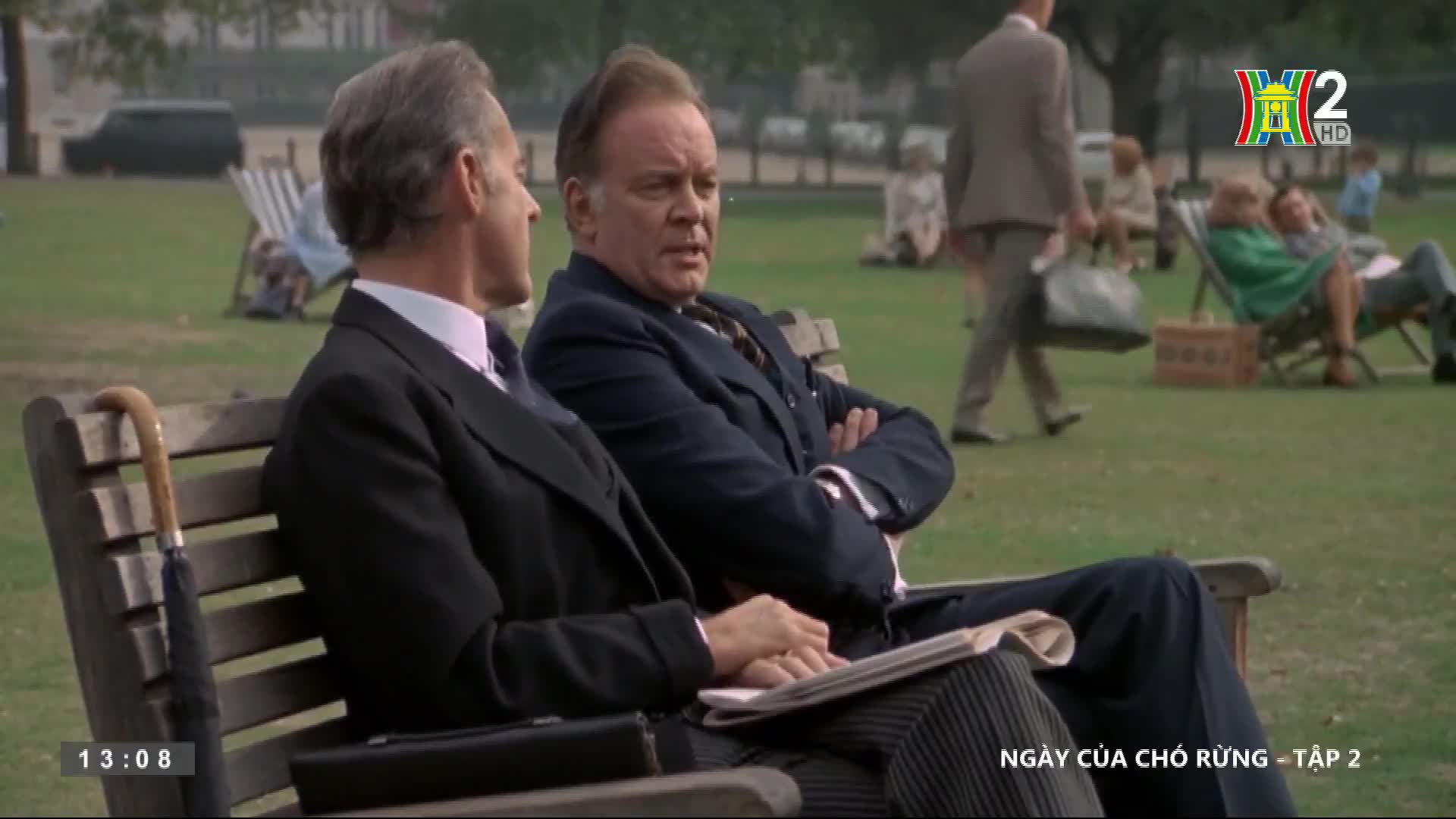 Two men in dark suits sit on a bench, one with arms crossed, the other holding a newspaper. People stroll across the green, and a couple relaxes in folding chairs; it's a scene from "Ngày Của Chó Rừng" on HanoiTV2.
