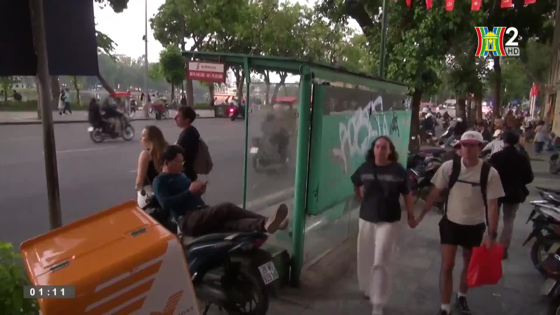 A couple strolls hand-in-hand down the sidewalk, while a man rests on a motorbike, scrolling on his phone. Motorbikes and pedestrians mingle along the street, under the watchful eye of HanoiTV2.

