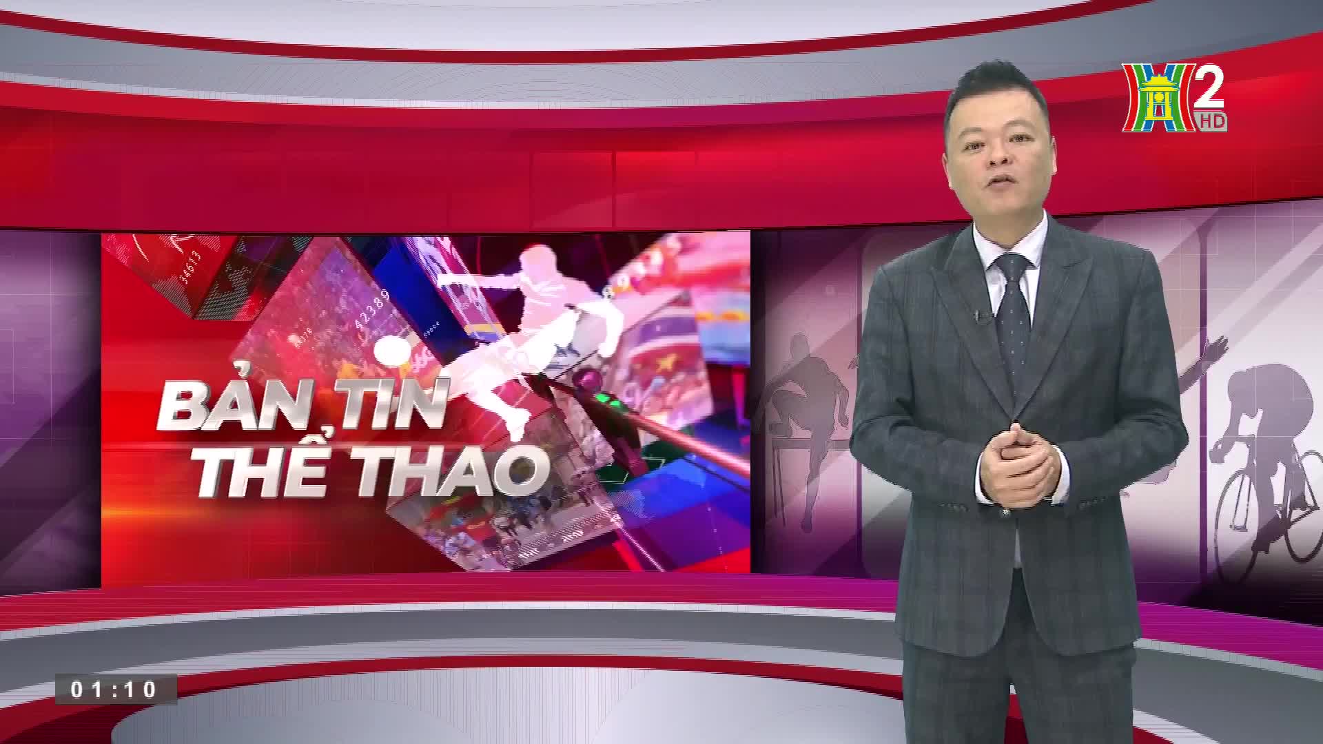 A man in a suit stands in front of a red and grey backdrop, speaking to the camera. The HanoiTV2 logo is visible in the top right corner, and the words "Bản Tin Thể Thao" are displayed on a screen behind him.
