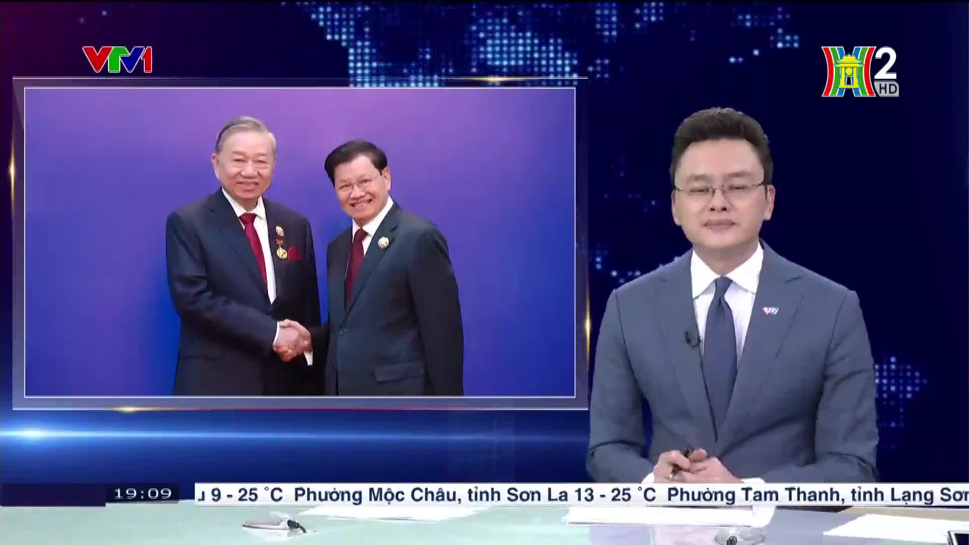 A framed image on HanoiTV2 shows two men shaking hands. Beside it, a news anchor in a suit looks directly at the camera.
