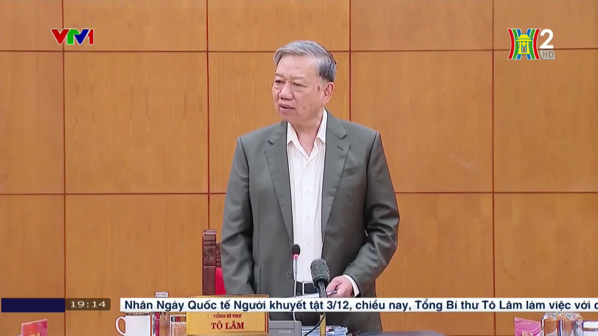 A man in a gray jacket speaks into a microphone, his gaze directed slightly to the left. The HanoiTV2 logo is visible in the top right corner, and the VTV1 logo is in the top left.
