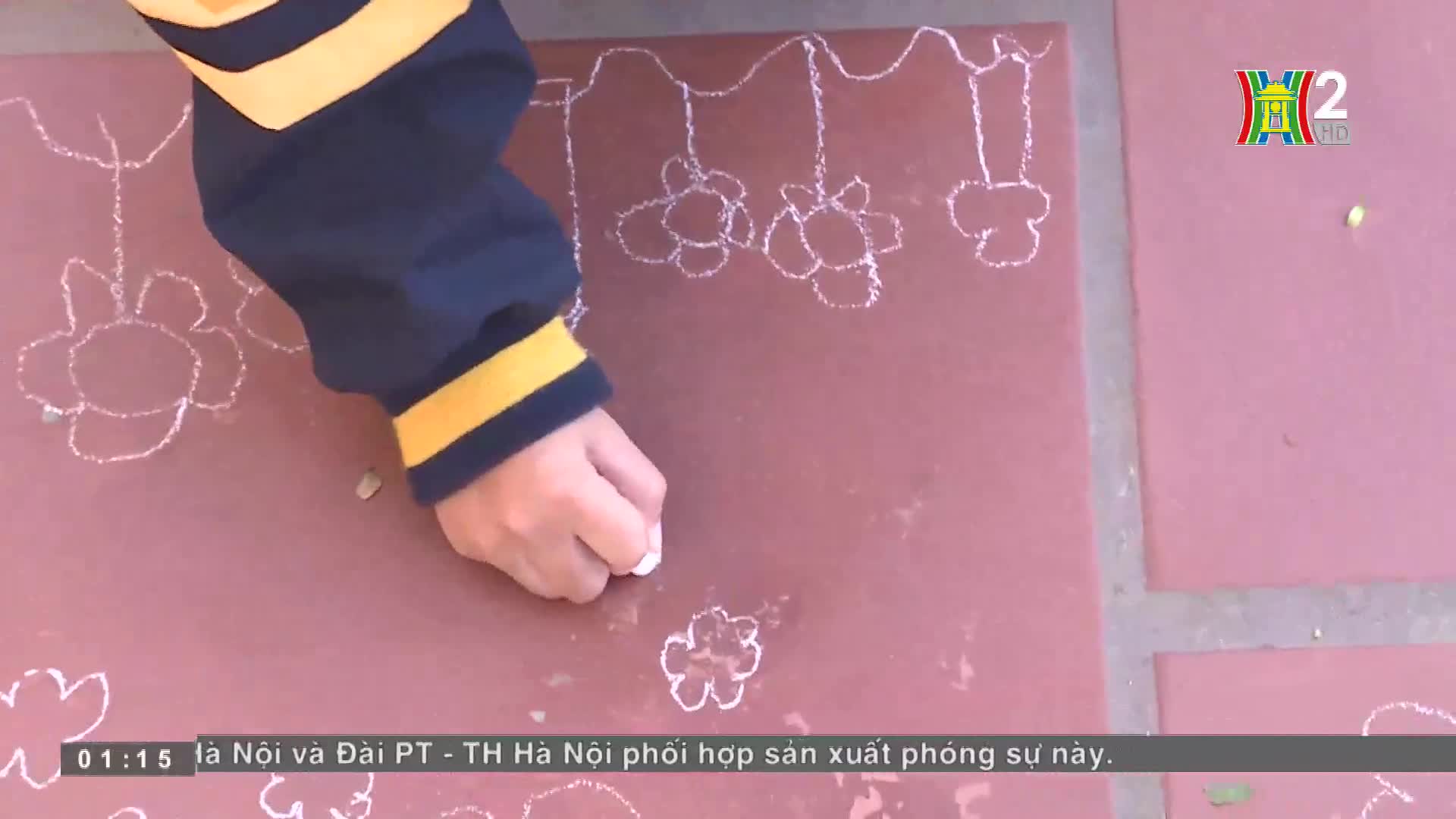 A hand, clad in a navy sleeve with yellow stripes, carefully draws white chalk flowers on a red surface. The HanoiTV2 logo is visible in the corner, and the Vietnamese caption scrolls along the bottom.

