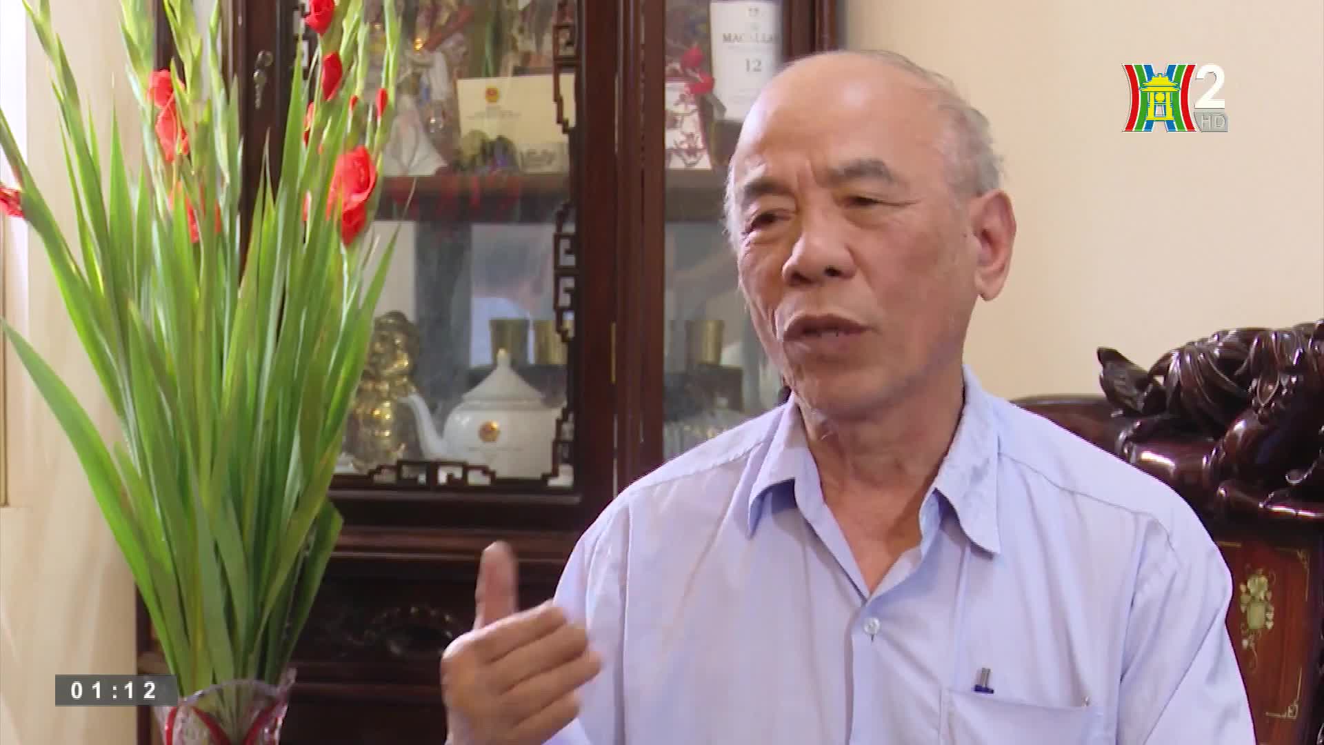A man in a light blue shirt gestures with his right hand as he speaks. Behind him, a vase of red gladioli stands next to a glass cabinet, and the HanoiTV2 logo is visible in the upper right corner.
