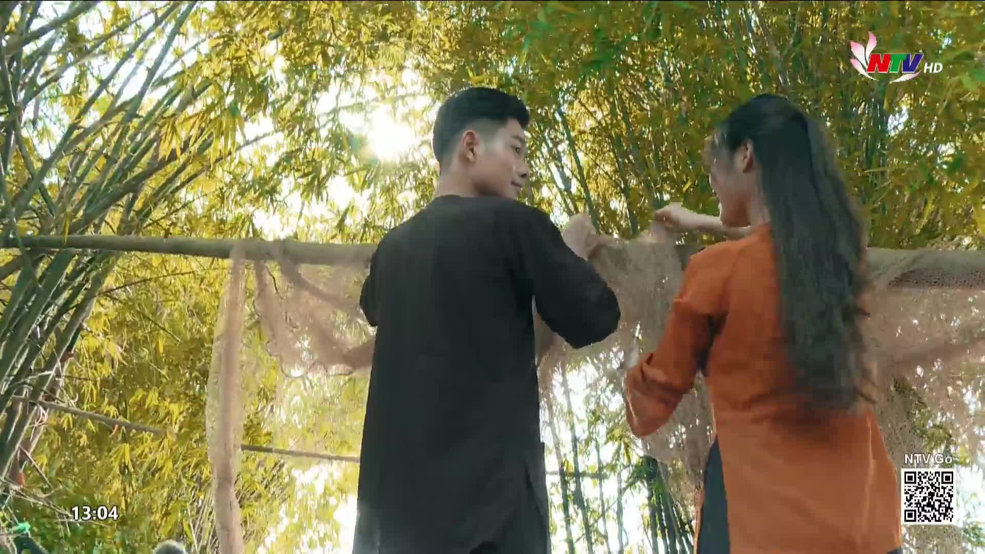 A young man and woman are carefully hanging a delicate, lacy net in front of a bamboo grove. The scene is bathed in sunlight, likely part of a broadcast from Nghe An TV in Vietnam.
