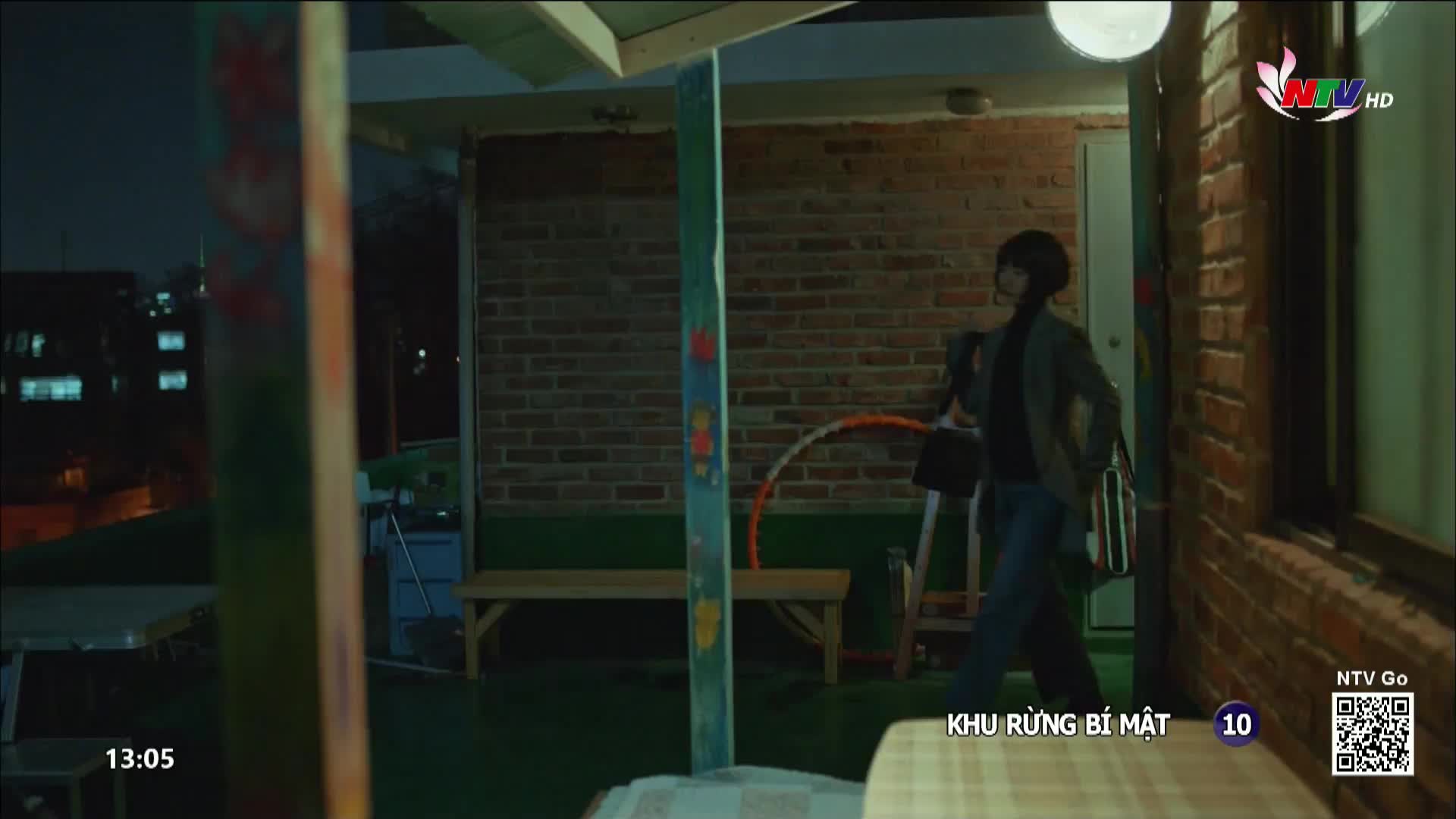 A person walks through a doorway, carrying a bag, as a scene from Nghe An TV plays on the screen. The time is 13:05, and the show "Khu Rừng Bí Mật" is on.
