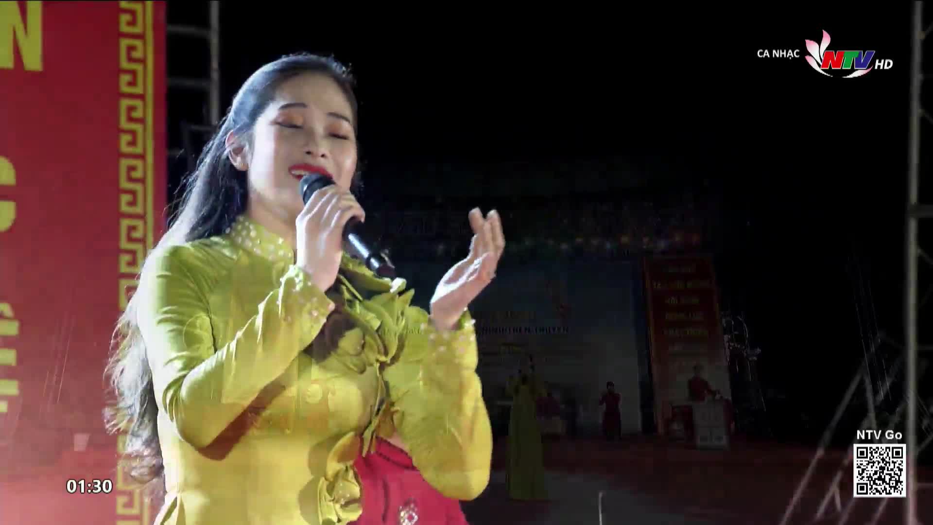 A woman in a bright yellow dress sings into a microphone, her hand gracefully gesturing as she performs. The stage is lit, and the Nghe An TV logo is visible in the corner.
