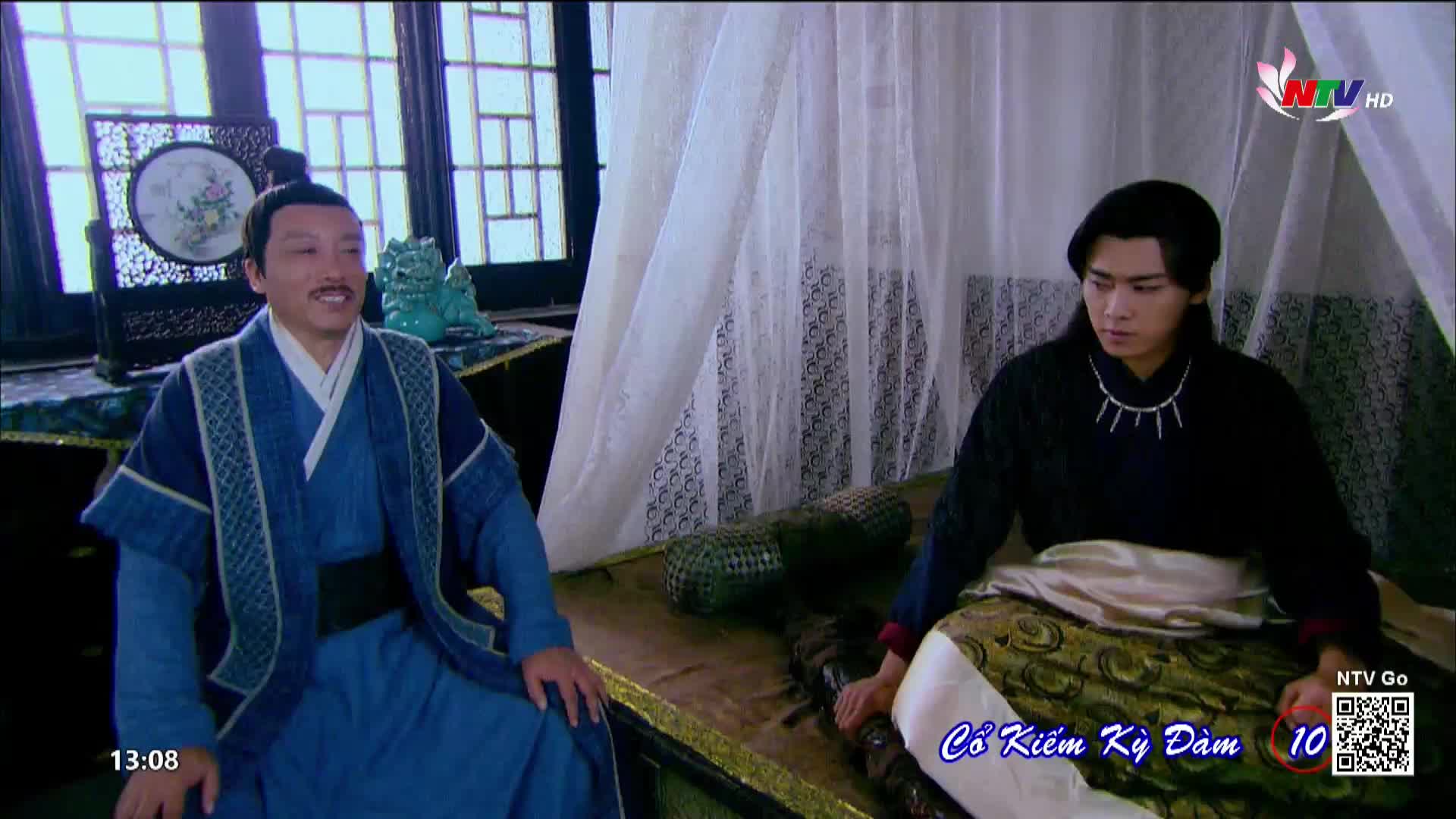 A man in blue robes smiles, seated on a low platform. Across from him, another man in black looks down, his hands resting on the cushions.
