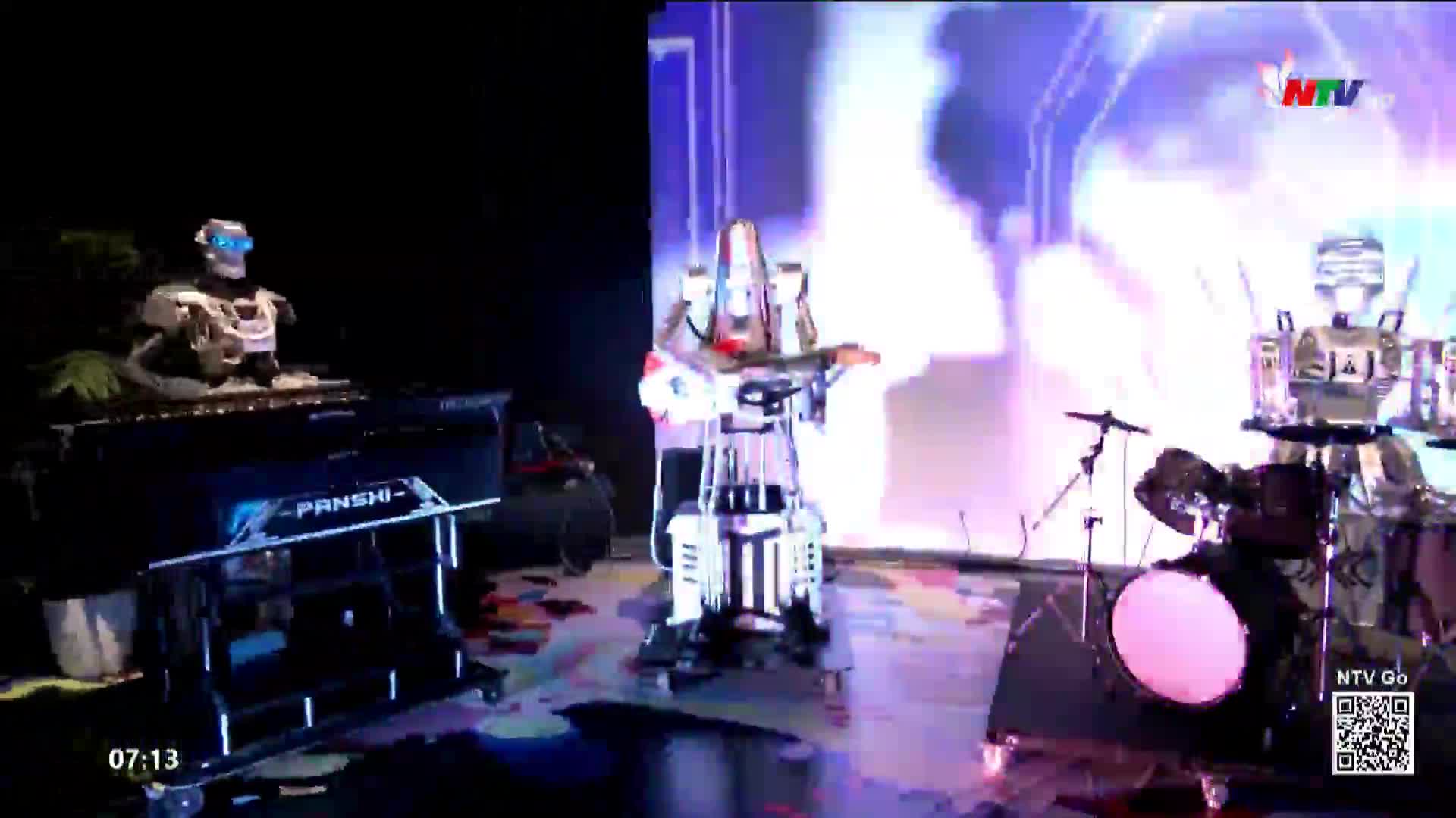 On the stage, three robots are performing. One plays a keyboard, another strums a guitar, and the third is behind a drum kit, all under the bright lights of Nghe An TV.
