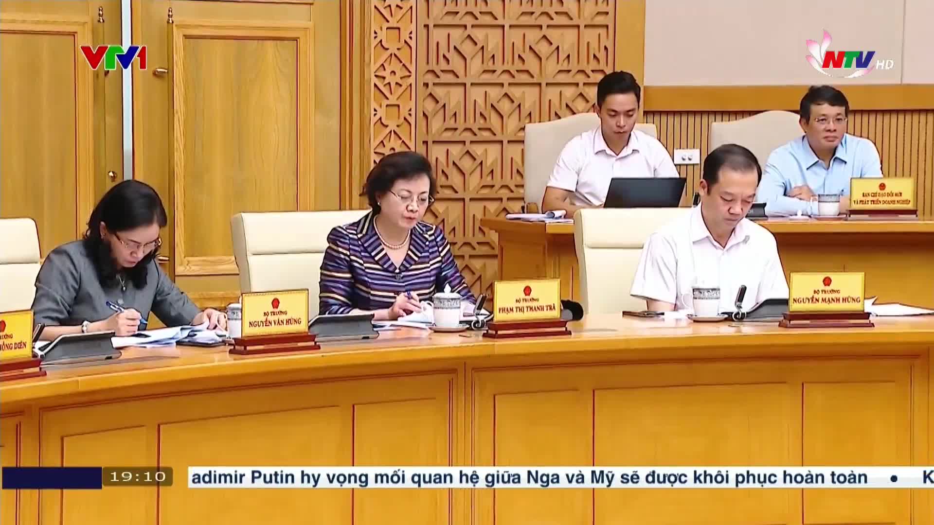 A group of people in a formal setting are seated around a large wooden table. The woman in the center is speaking, while others are looking at papers and taking notes.
