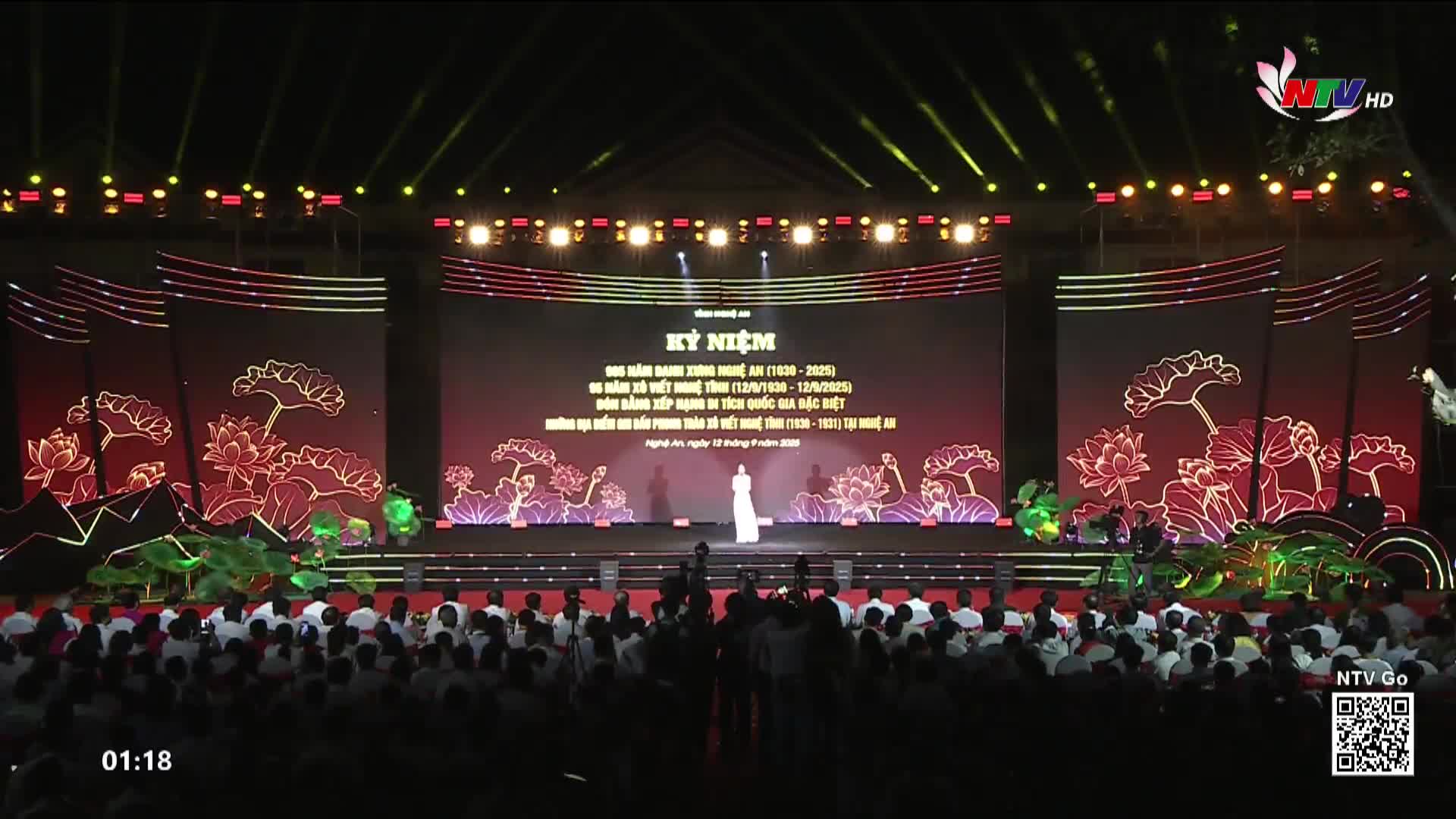 A woman in a long white dress stands centered on a stage, illuminated by spotlights, addressing a crowd. Behind her, large screens display text and floral designs, all part of the Nghe An TV broadcast.
