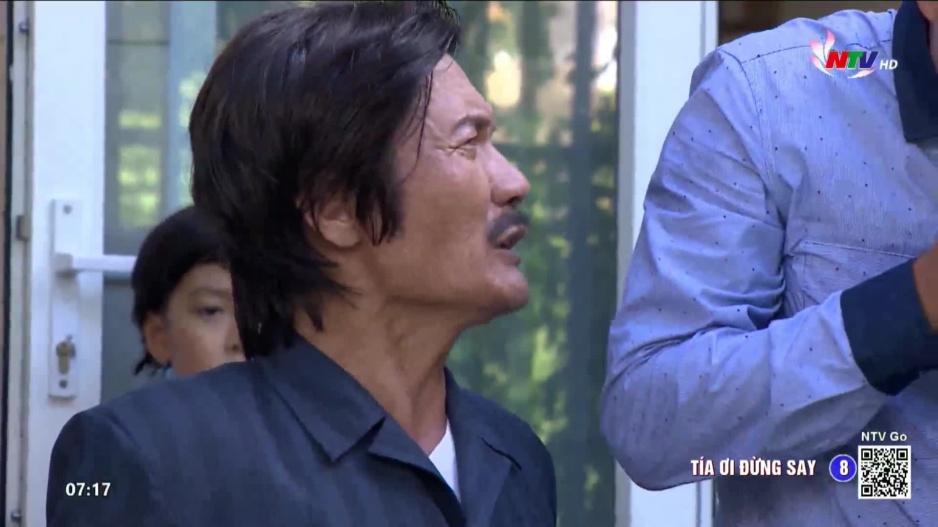 The man in the dark jacket is talking, his mouth open in mid-sentence. Behind him, a child peers out from behind a door, and the Nghe An TV logo is visible in the corner.
