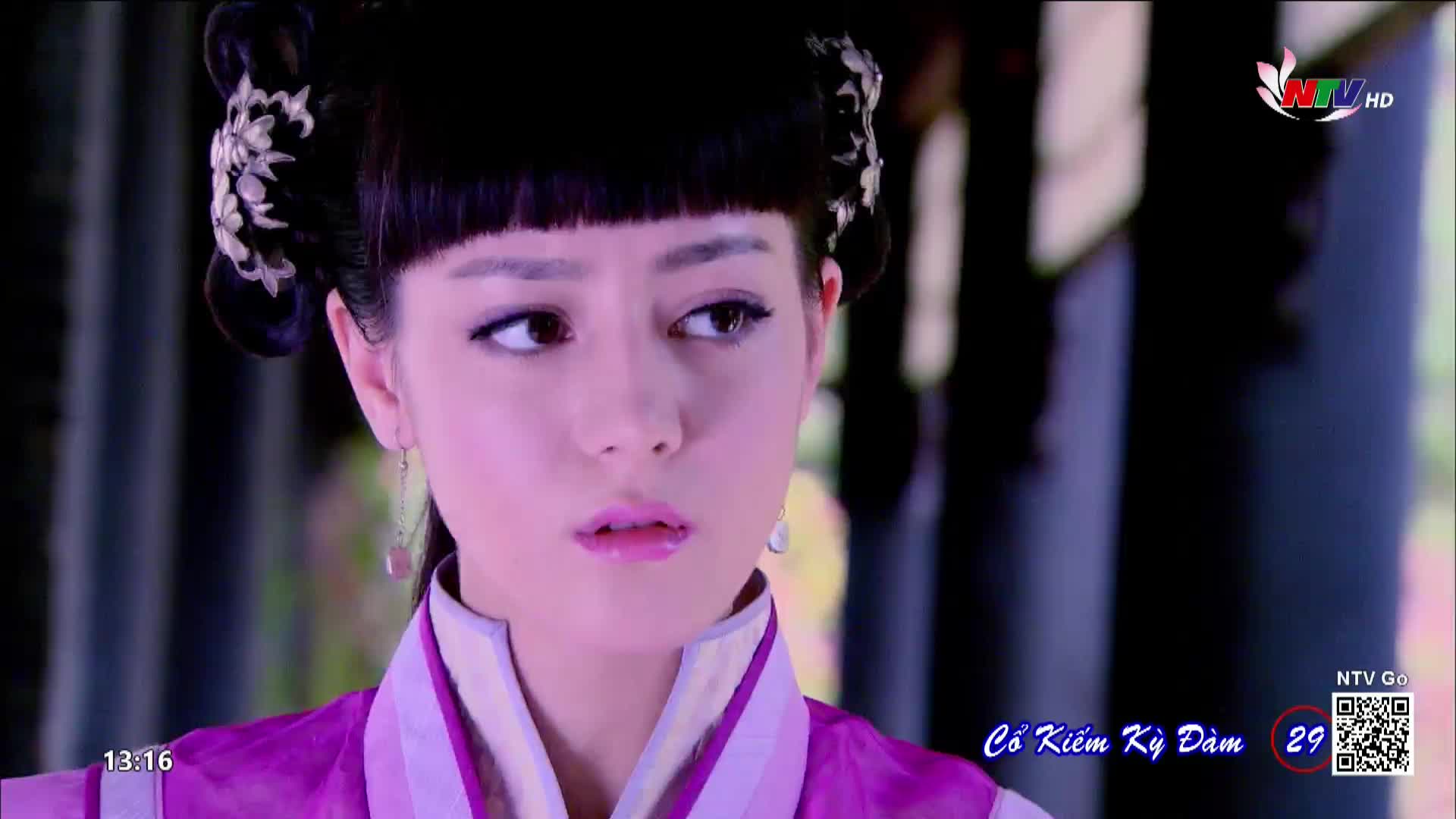 The woman's eyes dart to the side, her lips slightly parted as if she's about to speak.  On Nghe An TV, the scene from "Cổ Kiếm Kỳ Đàm" is paused, with a timestamp in the corner.
