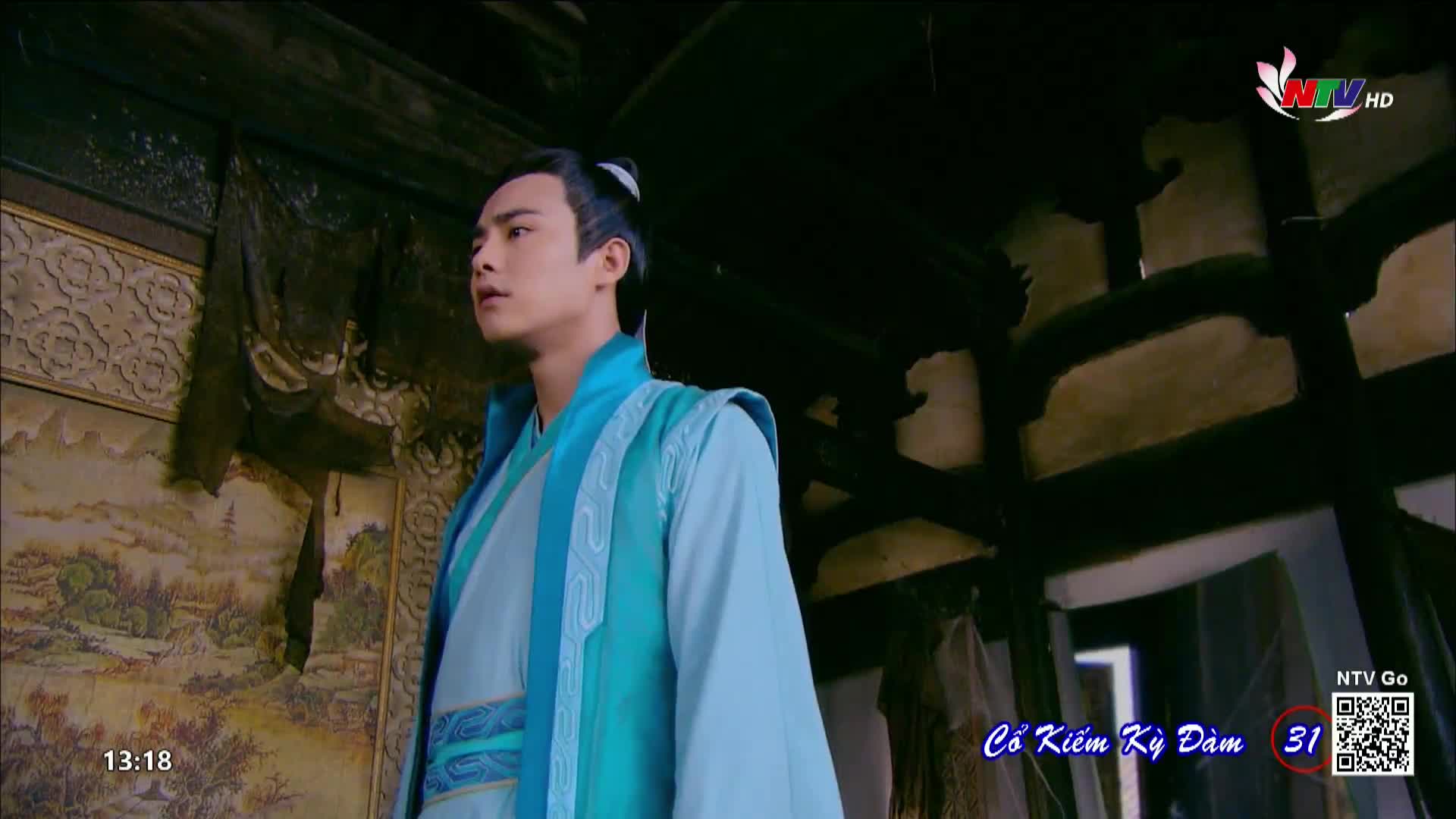 A young man in a light blue robe stands in a dimly lit room, his gaze fixed on something out of frame. The scene appears to be from a Vietnamese television show, as indicated by the Nghe An TV logo in the corner.
