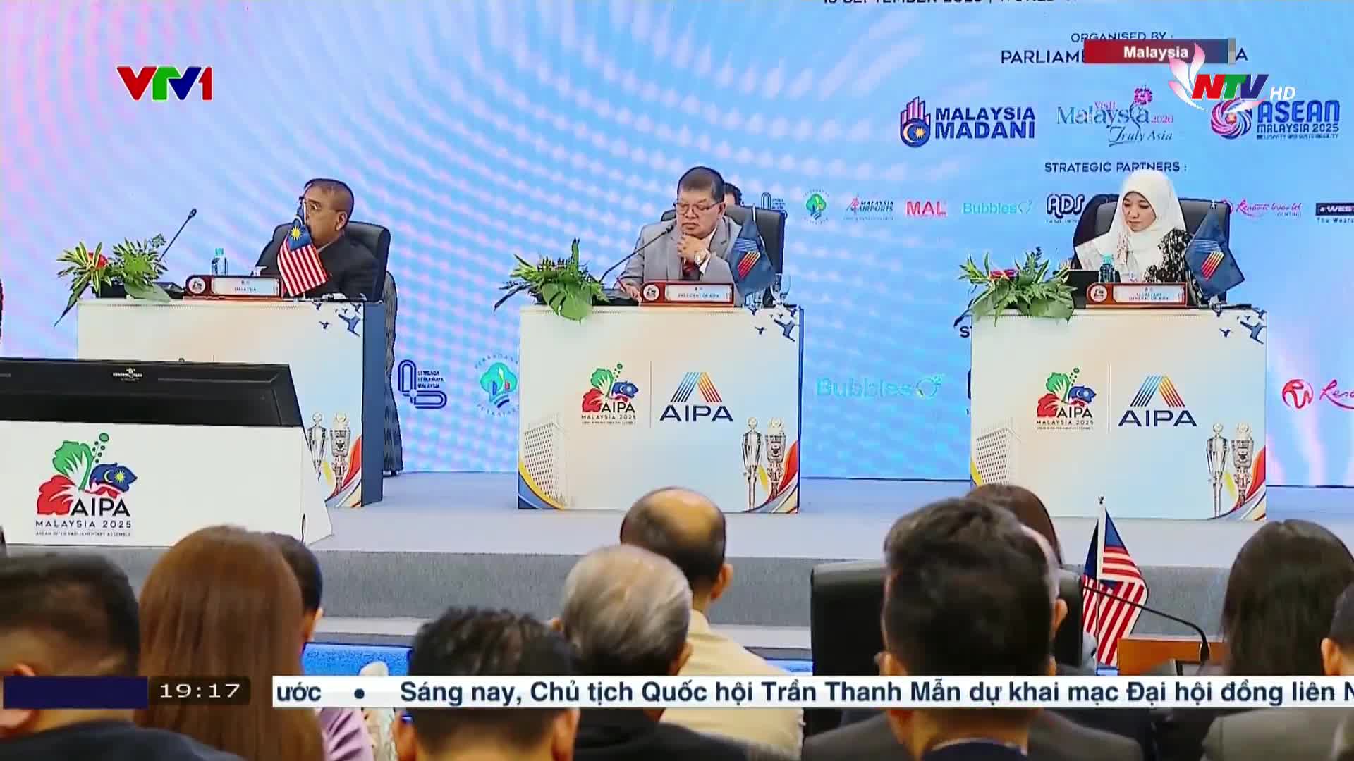 Three people sit at a table, each with a small Malaysian flag, facing an audience. The event, broadcast on Nghe An TV, is clearly a conference, with the AIPA logo displayed prominently.
