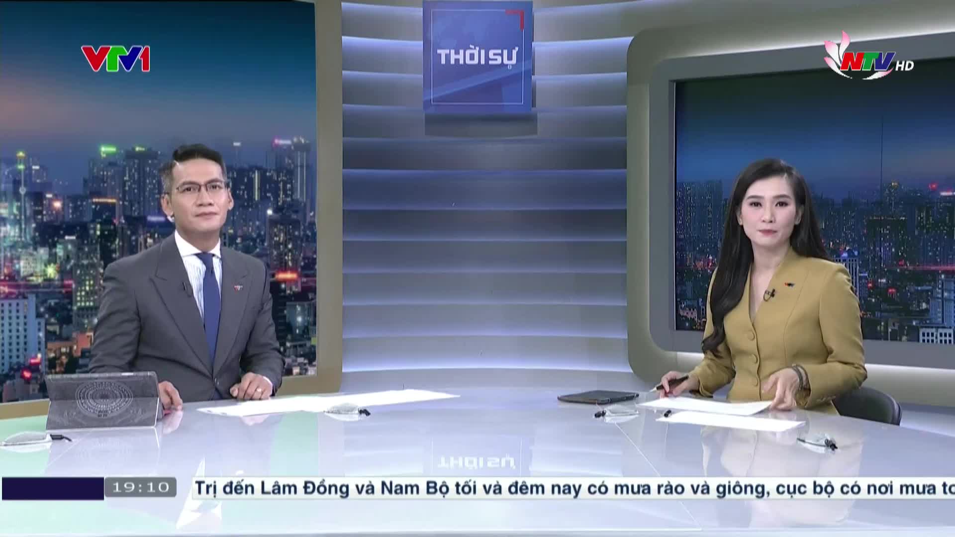 Two news anchors sit at a desk, facing the camera in a studio. The man wears a suit and tie, while the woman is in a yellow blazer; both are poised and ready to speak.
