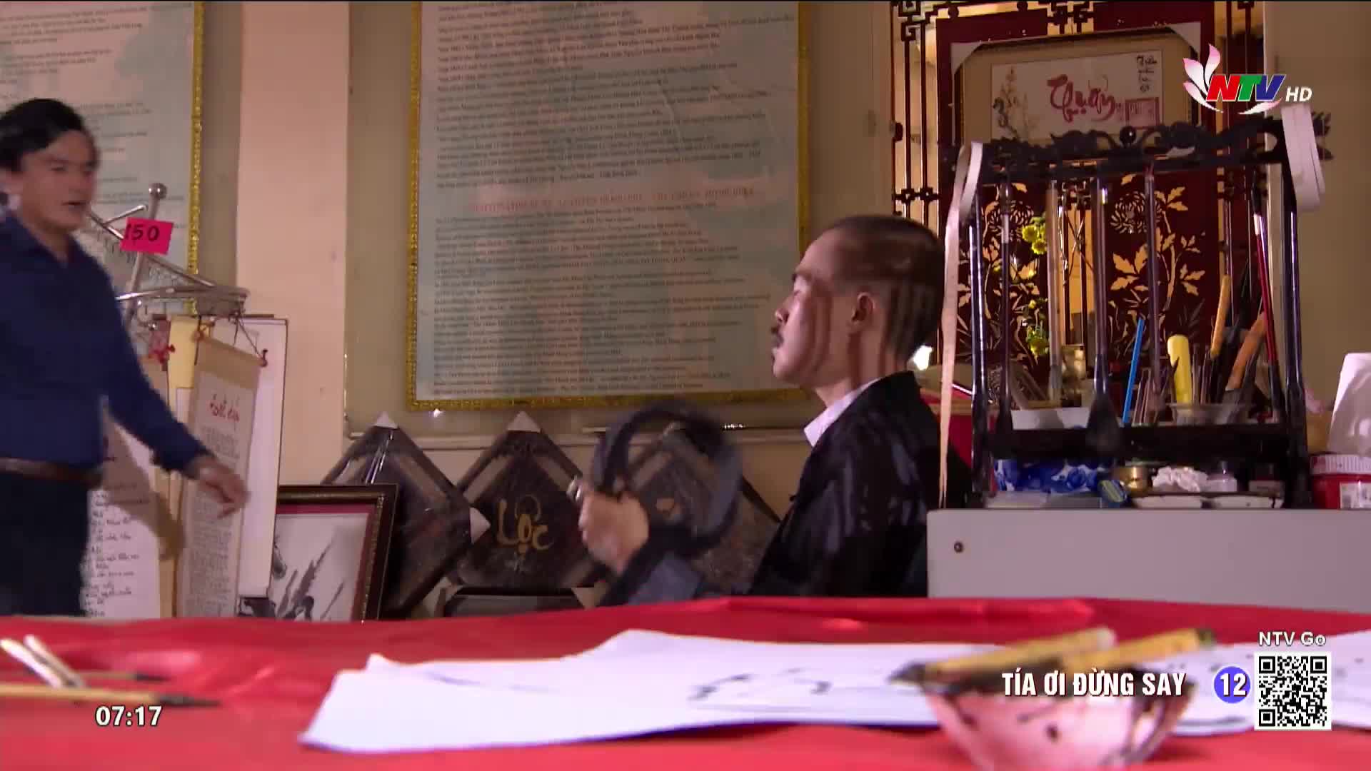 A man in a blue shirt gestures emphatically toward the bald-headed man seated behind a red-covered table. The seated man holds a fan, his face illuminated by the light filtering through the window, as the Nghe An TV logo appears in the corner.
