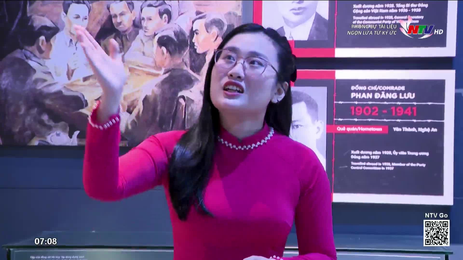 A woman in a bright pink áo dài gestures toward a painting and information panels, speaking animatedly. The Nghe An TV logo is visible in the corner, and the time is just after seven in the morning.
A woman in a bright pink áo dài gestures toward a painting and information panels, speaking animatedly. The Nghe An TV logo is visible in the corner, and the time is just after seven in the morning.