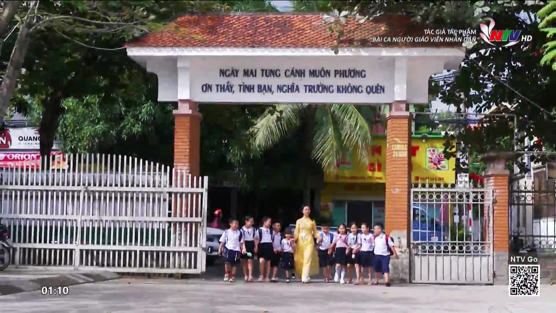 A group of children in school uniforms walk toward the camera, escorted by a woman in a yellow dress. The scene is broadcast on Nghe An TV, with the school's gate and a sign in Vietnamese visible.
