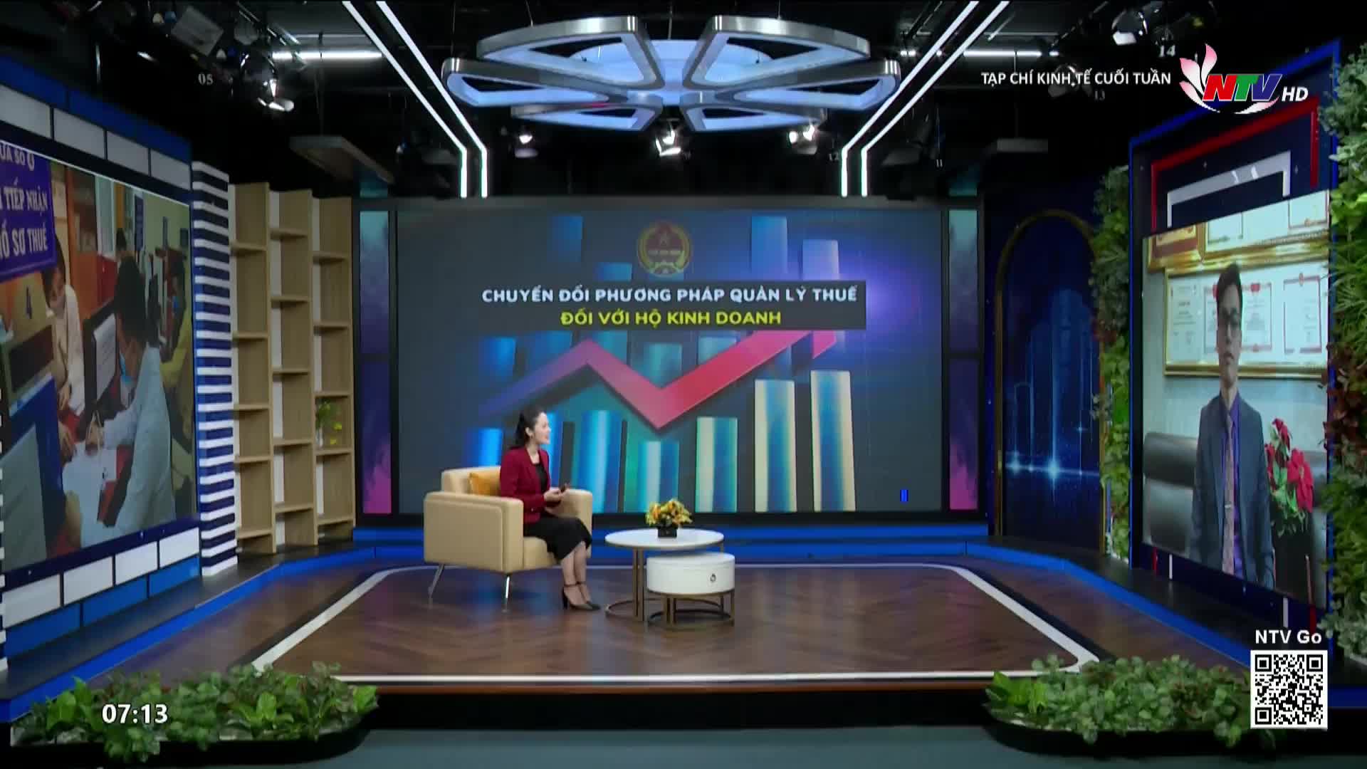 In a brightly lit studio, a woman in a red jacket sits in a beige armchair, facing a large screen. Behind her, the screen displays a title about tax management in Vietnamese, with a rising graph.
