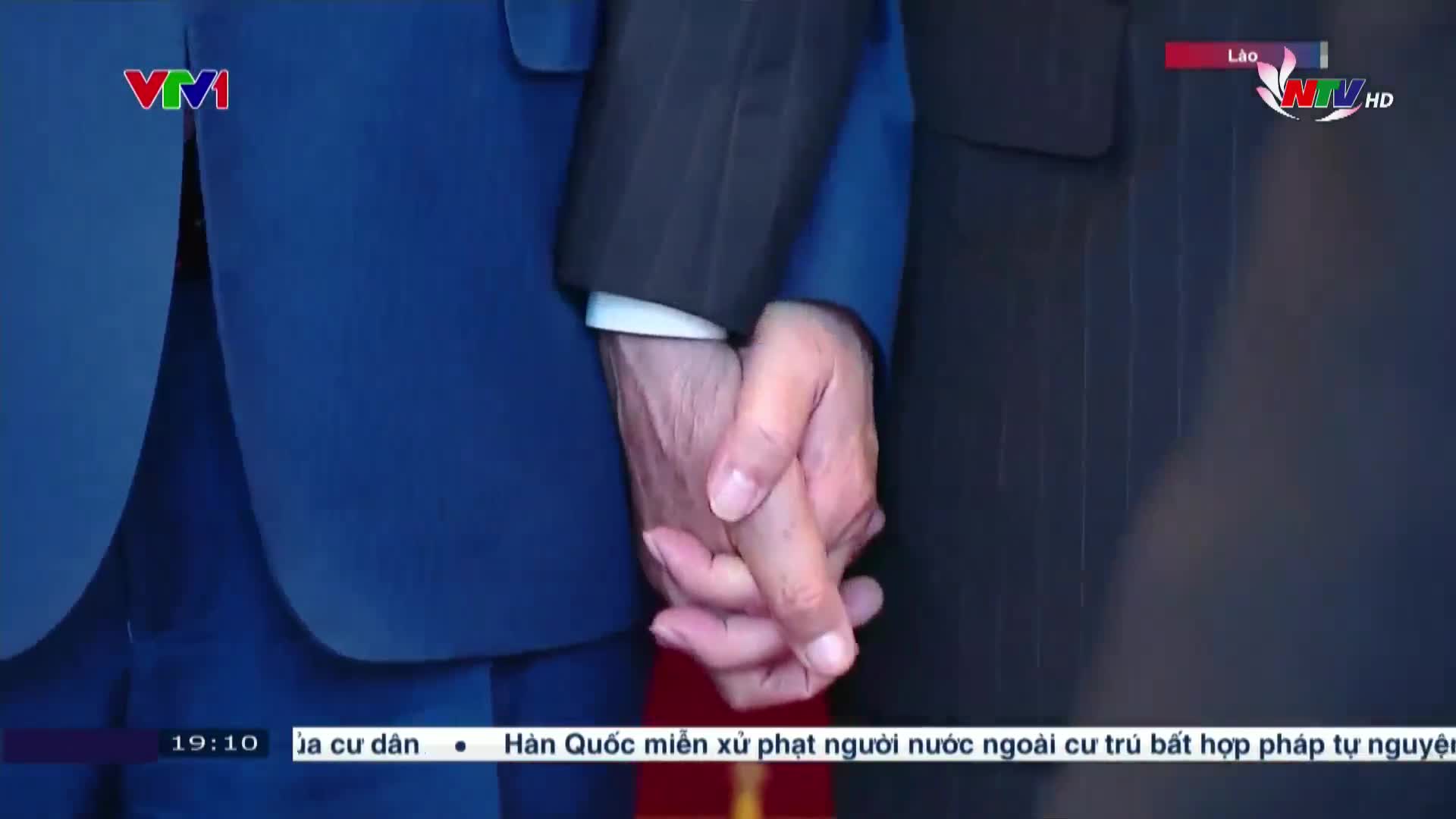 Two men's hands are clasped tightly together, one wearing a dark suit, the other a blue blazer. The scene is broadcast on Nghe An TV, with a VTV logo visible in the corner.
