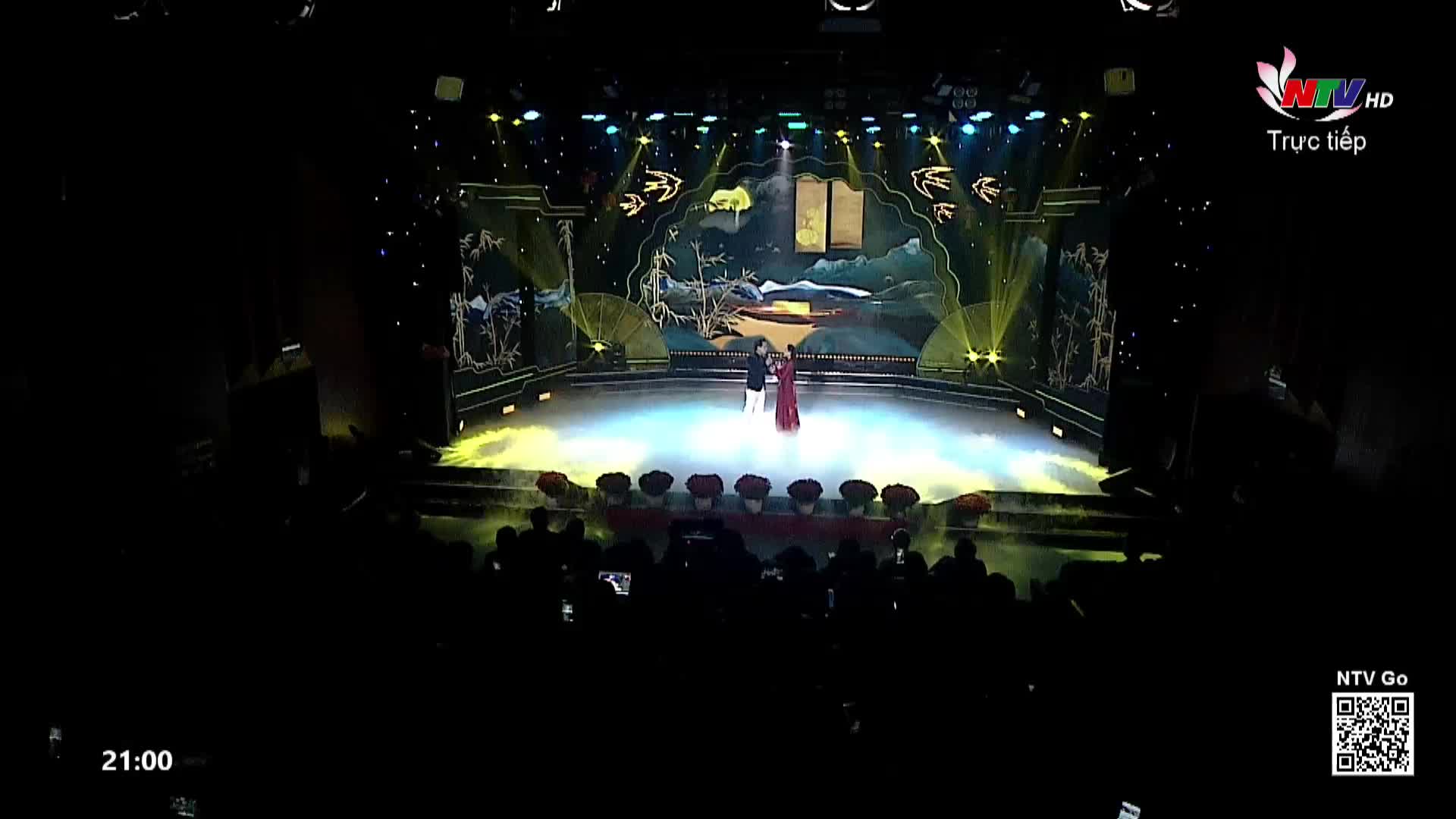 Two performers stand center stage, bathed in a bright spotlight. The backdrop depicts a serene landscape, reminiscent of traditional Vietnamese art.