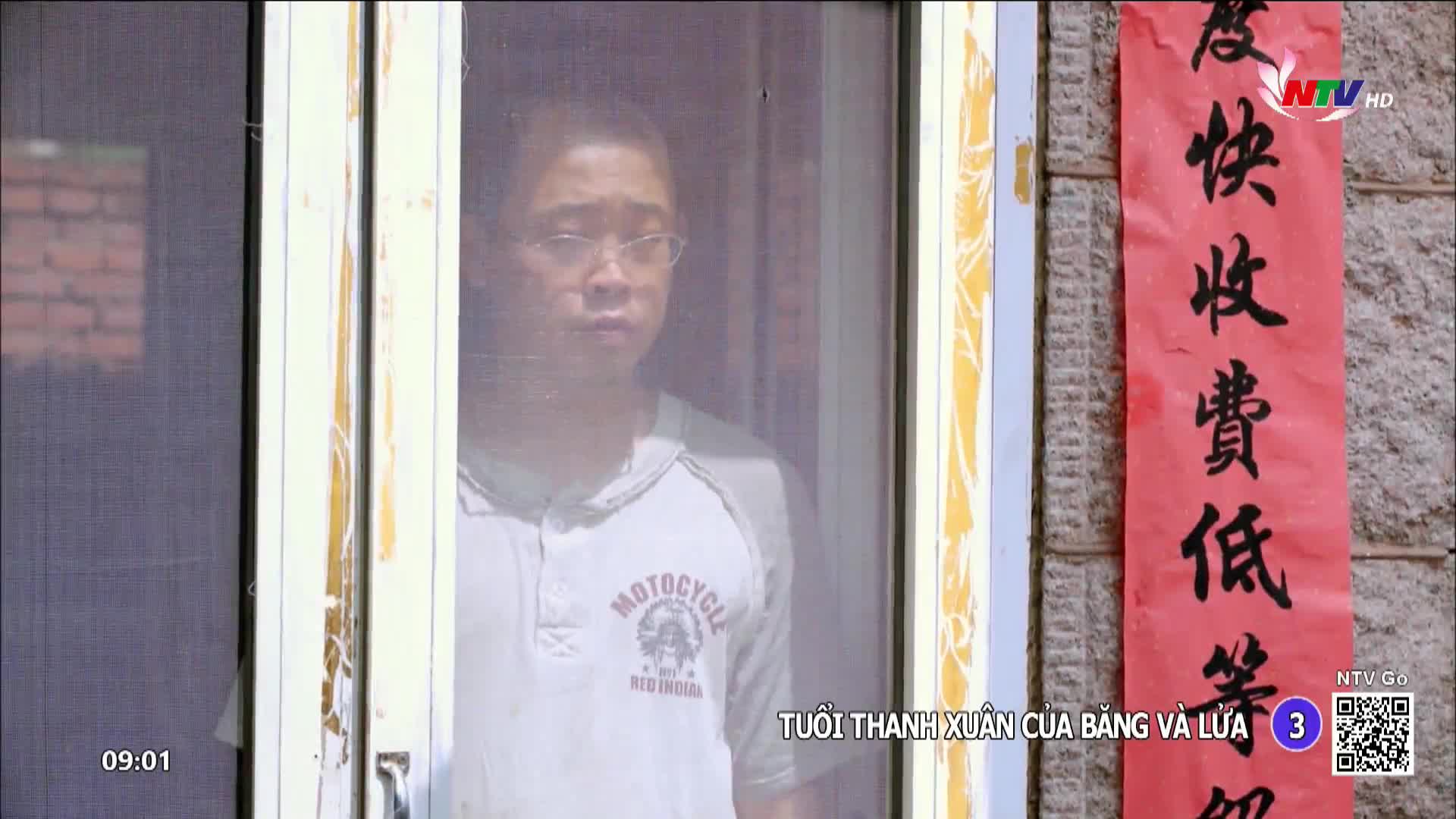 A young man with glasses looks out from behind a screen door. A red banner with black calligraphy hangs on the wall beside him.