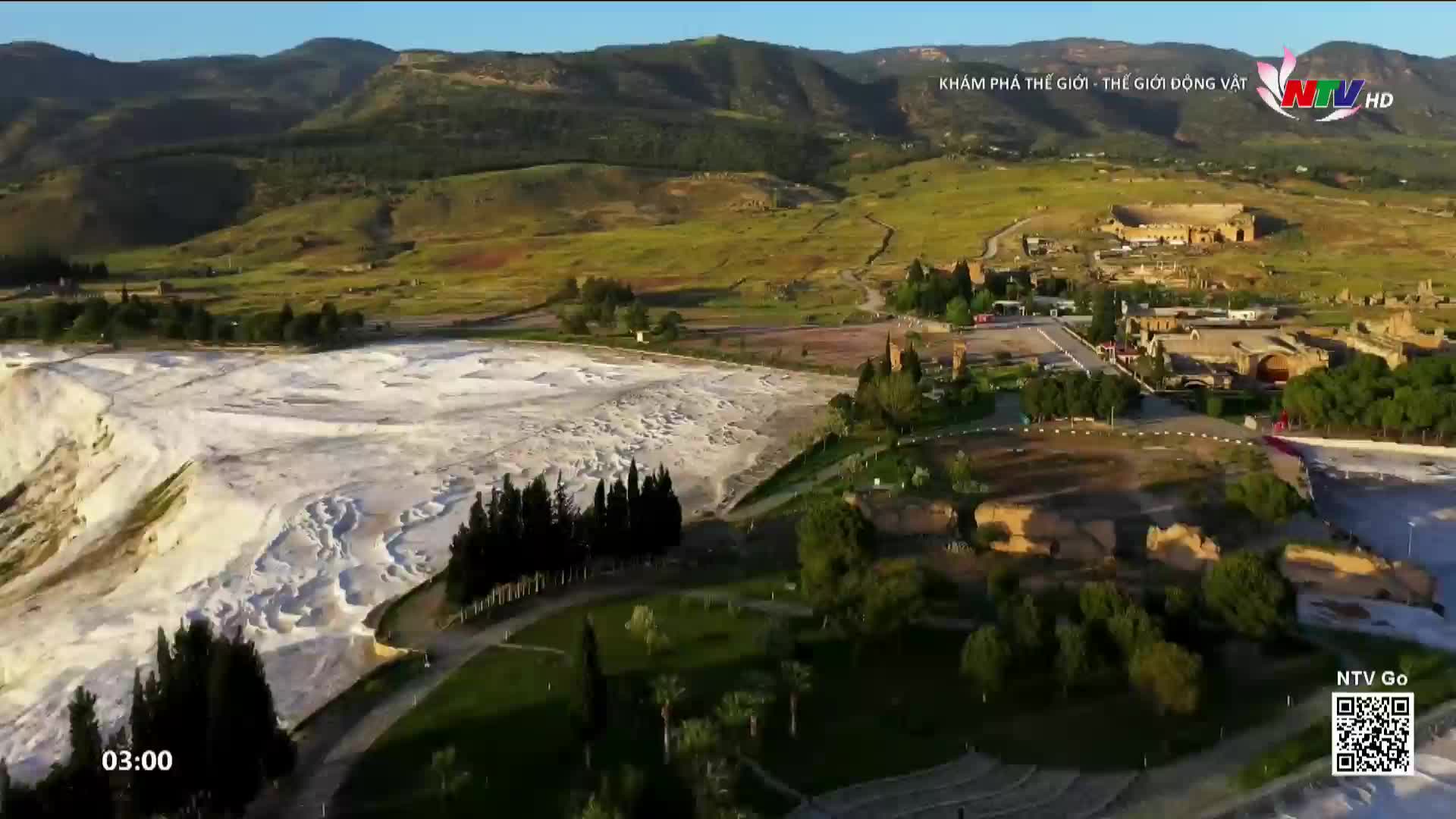 The white terraces of Pamukkale stretch out before me, a stark contrast to the green hills and ancient ruins. A few cars navigate the road leading to the historic site, their headlights glinting in the afternoon sun.