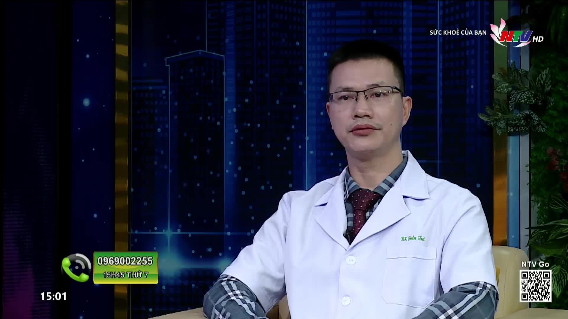 A doctor in a white coat sits on a chair, facing forward. Behind him, a digital cityscape glows on a screen, with the Nghe An TV logo visible.