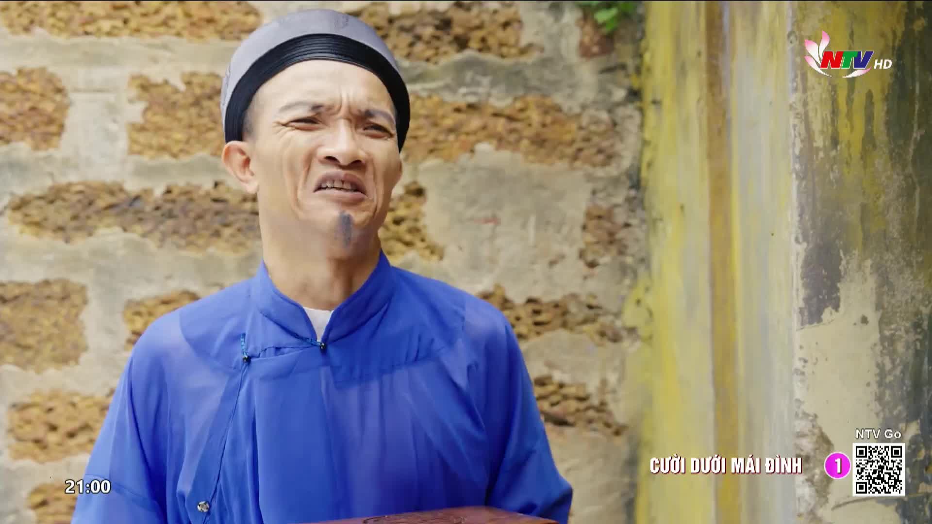 A man in a blue robe and traditional hat makes a pained expression. He holds a small, dark box in his hands.